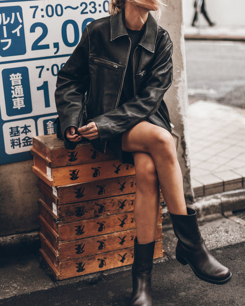 Person wearing a black leather jacket and boots sitting on wooden boxes against a street background.