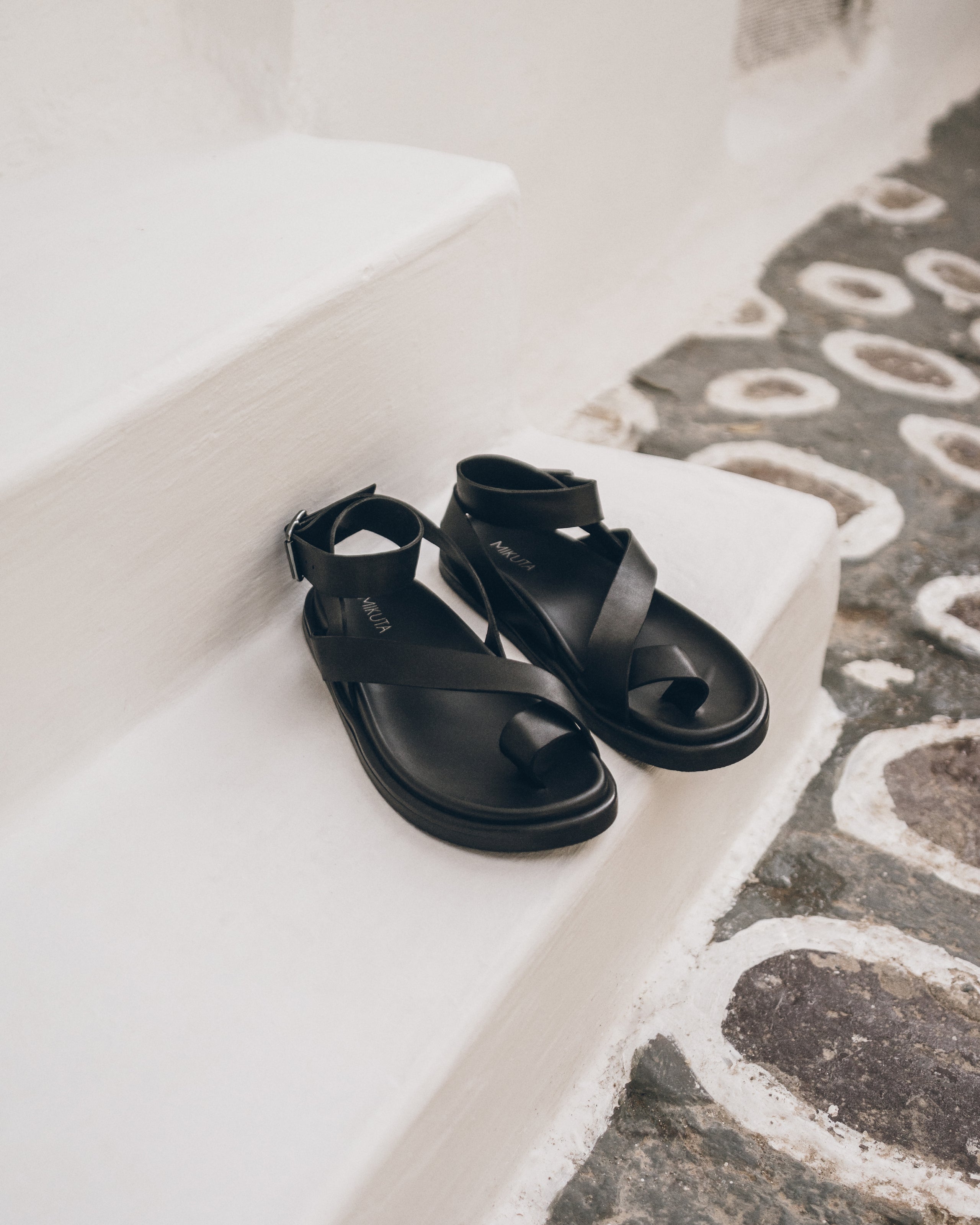 Black leather sandals on a white ledge with a patterned floor.