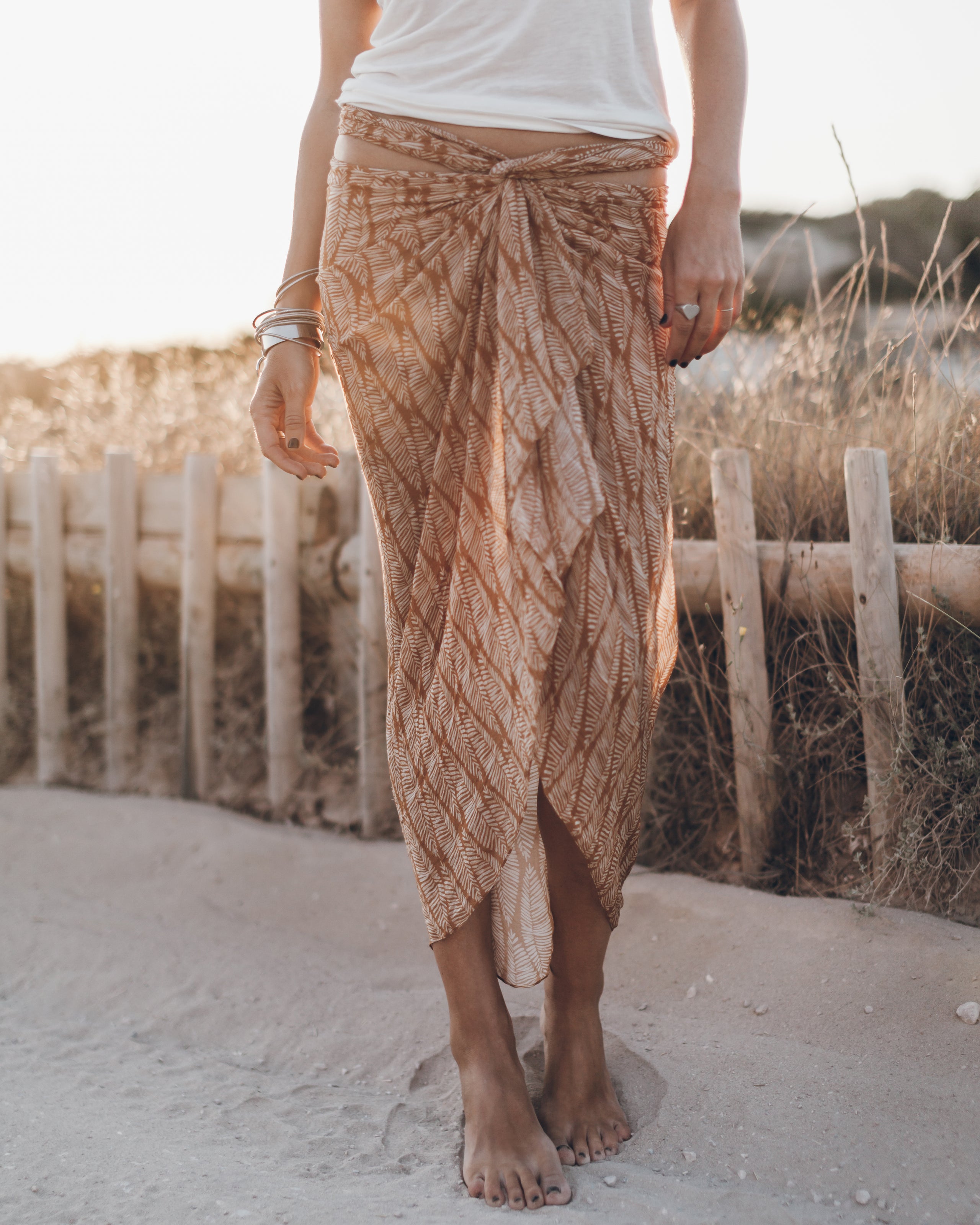 Person wearing a patterned skirt standing on a sandy path with a wooden fence and grassy area in the background.
