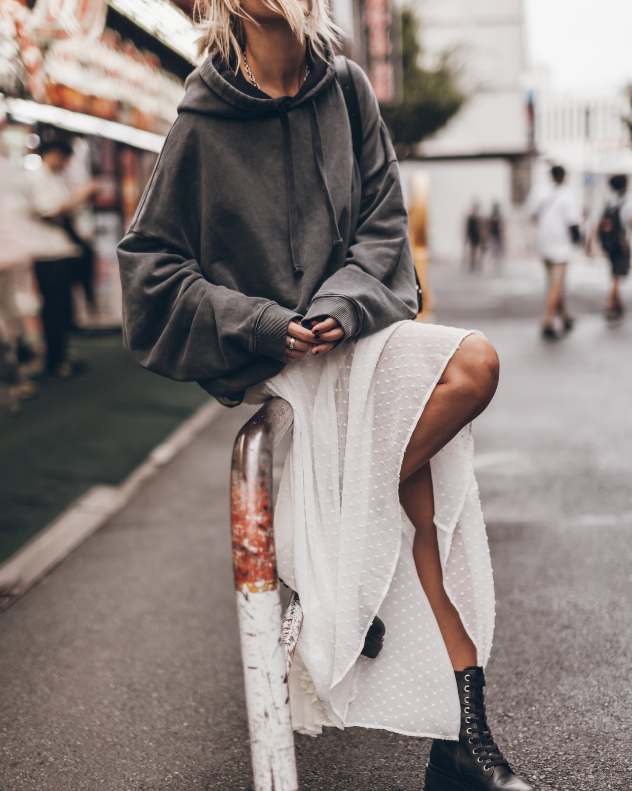 Person wearing a gray oversized hoodie and white skirt sitting on a metal railing outdoors.