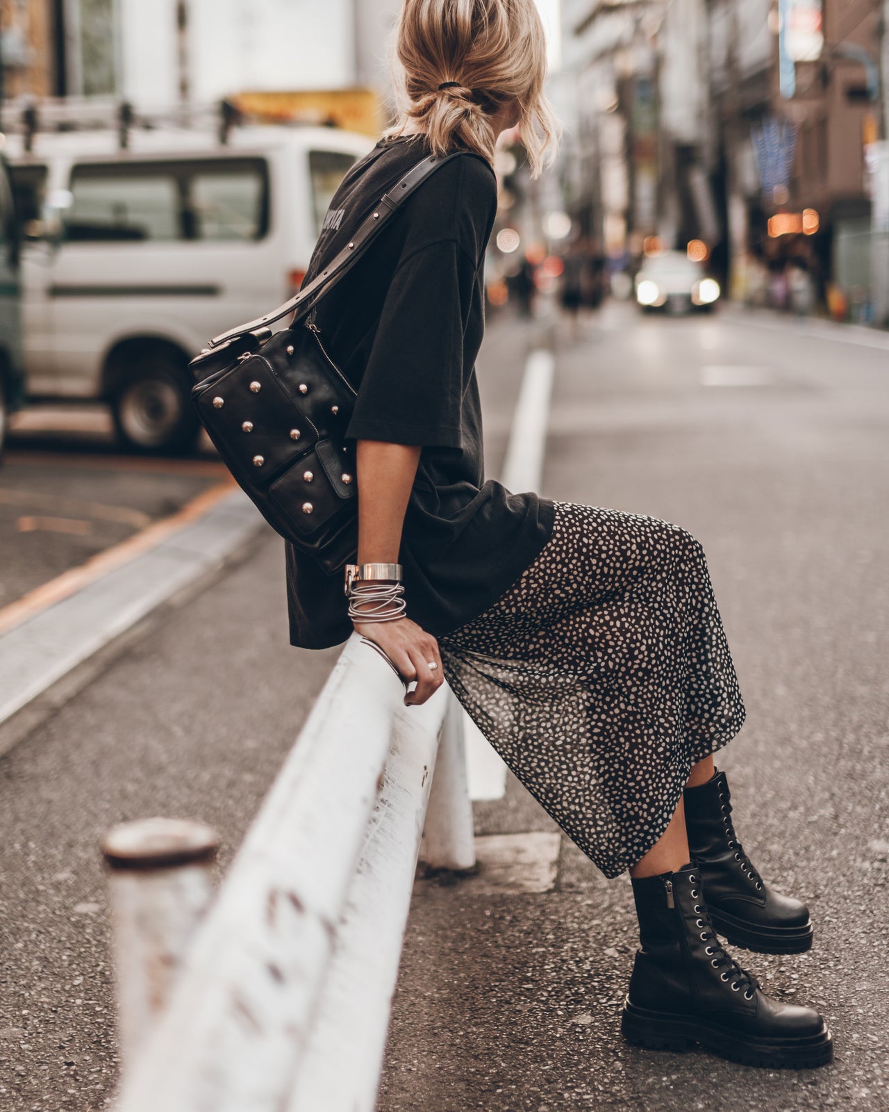 Woman sitting on a street curb wearing a black jacket, patterned skirt, and black boots.