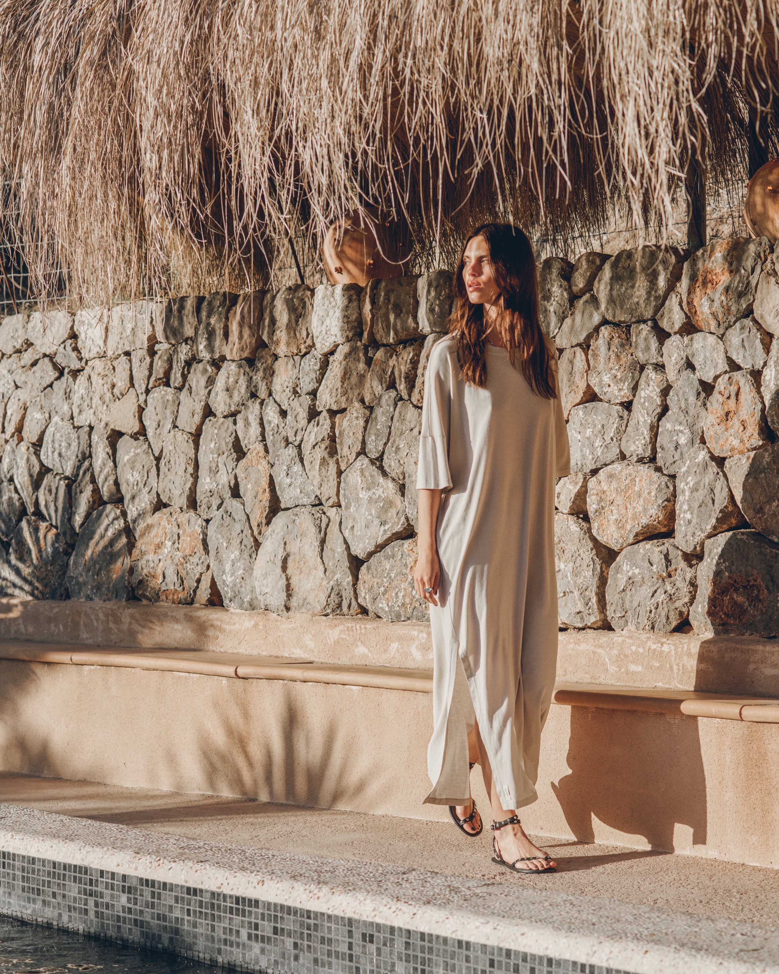 Woman in a white dress standing by a stone wall and poolside with a thatched roof in the background.