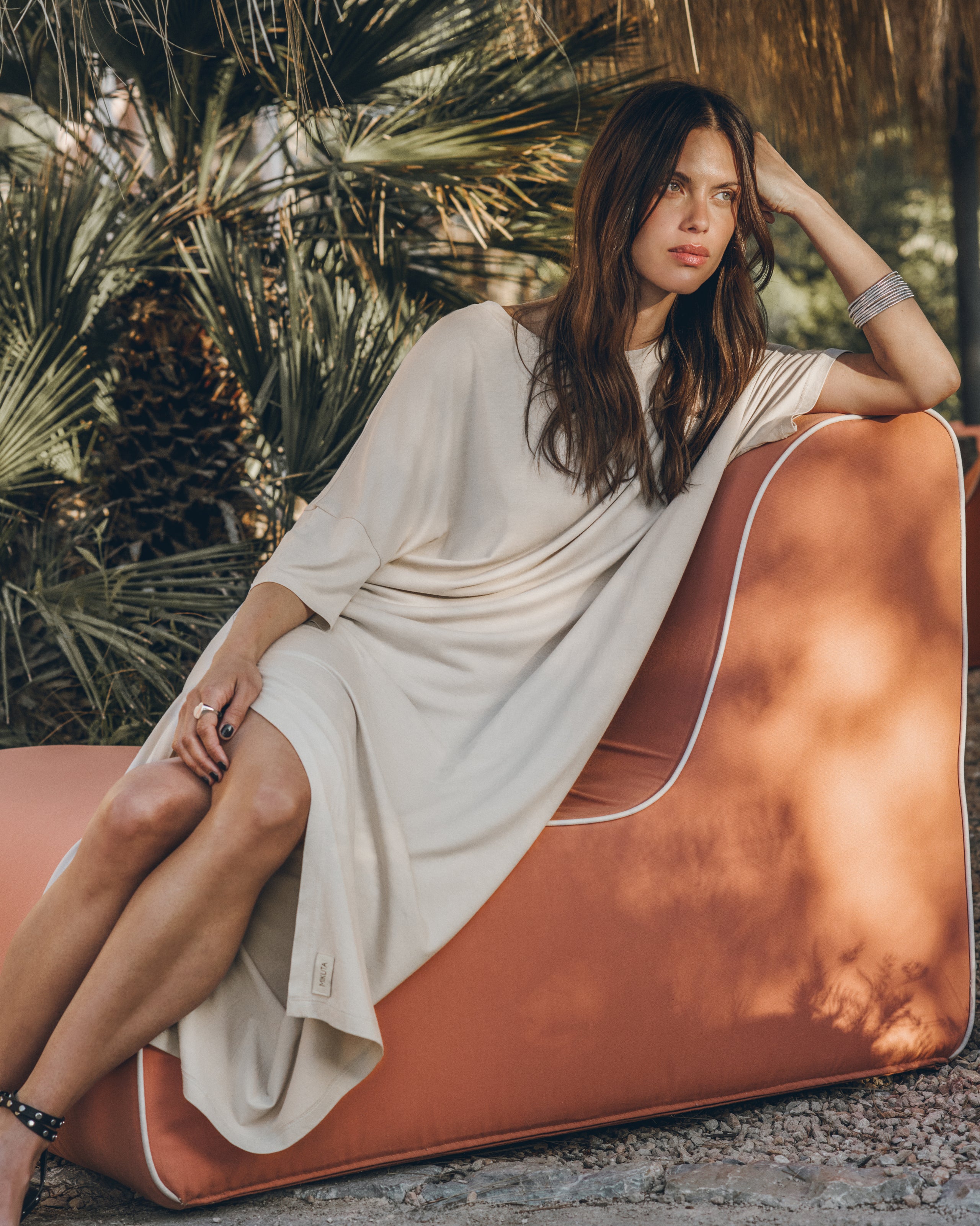 Woman in a white dress sitting on a brown chair with palm leaves in the background