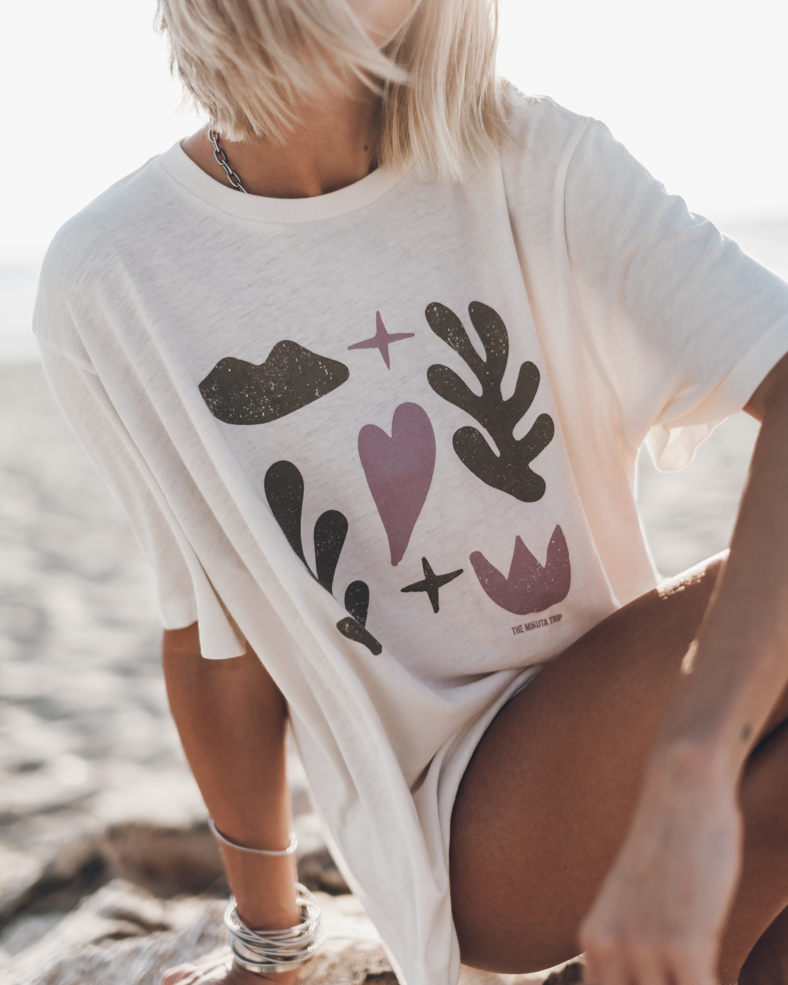 Woman wearing a cream-colored graphic tee with a silver chain necklace and stacked bracelets, sitting on a sandy surface.