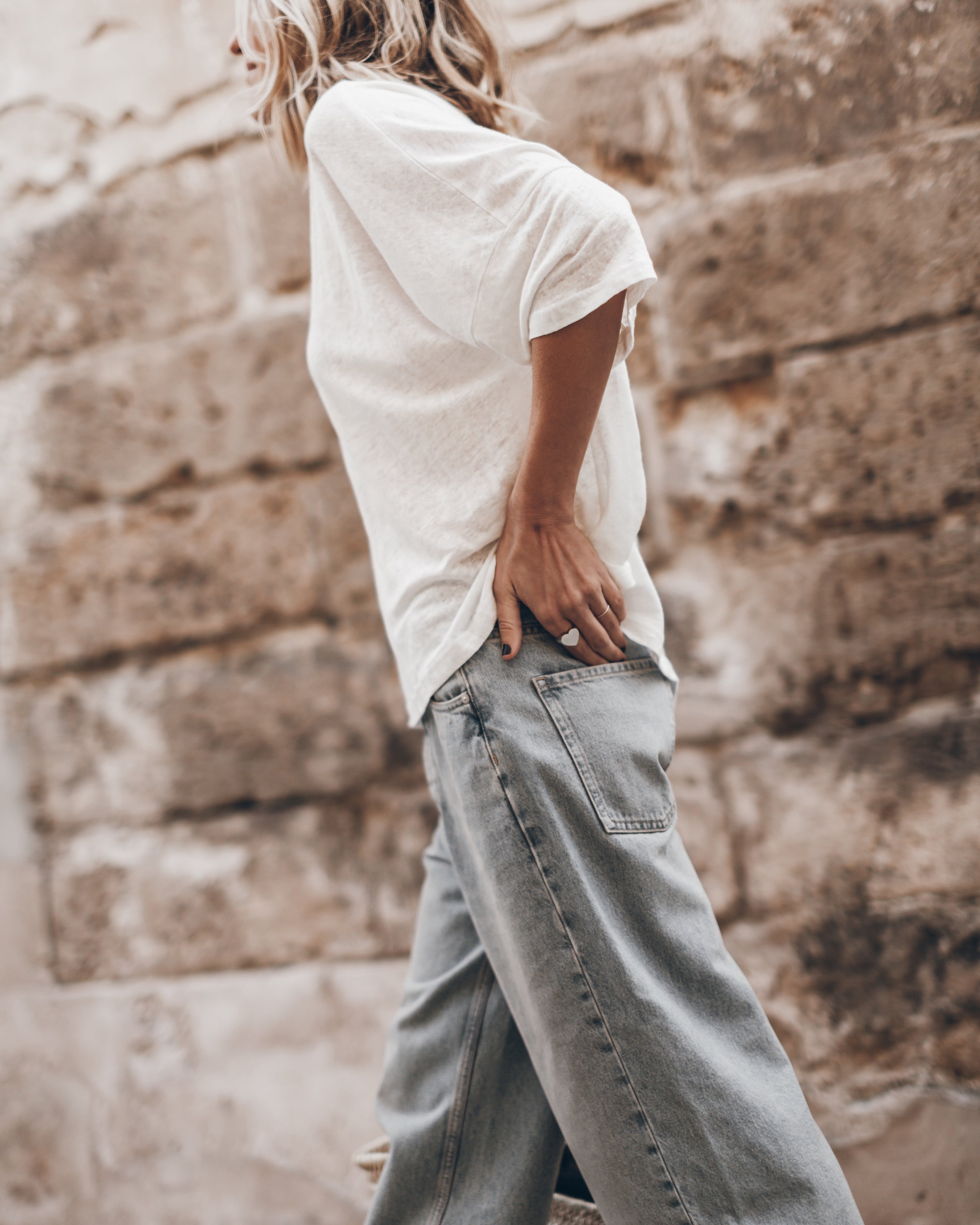 White loose short-sleeve linen t-shirt paired with light blue wide-leg jeans against a stone wall background.