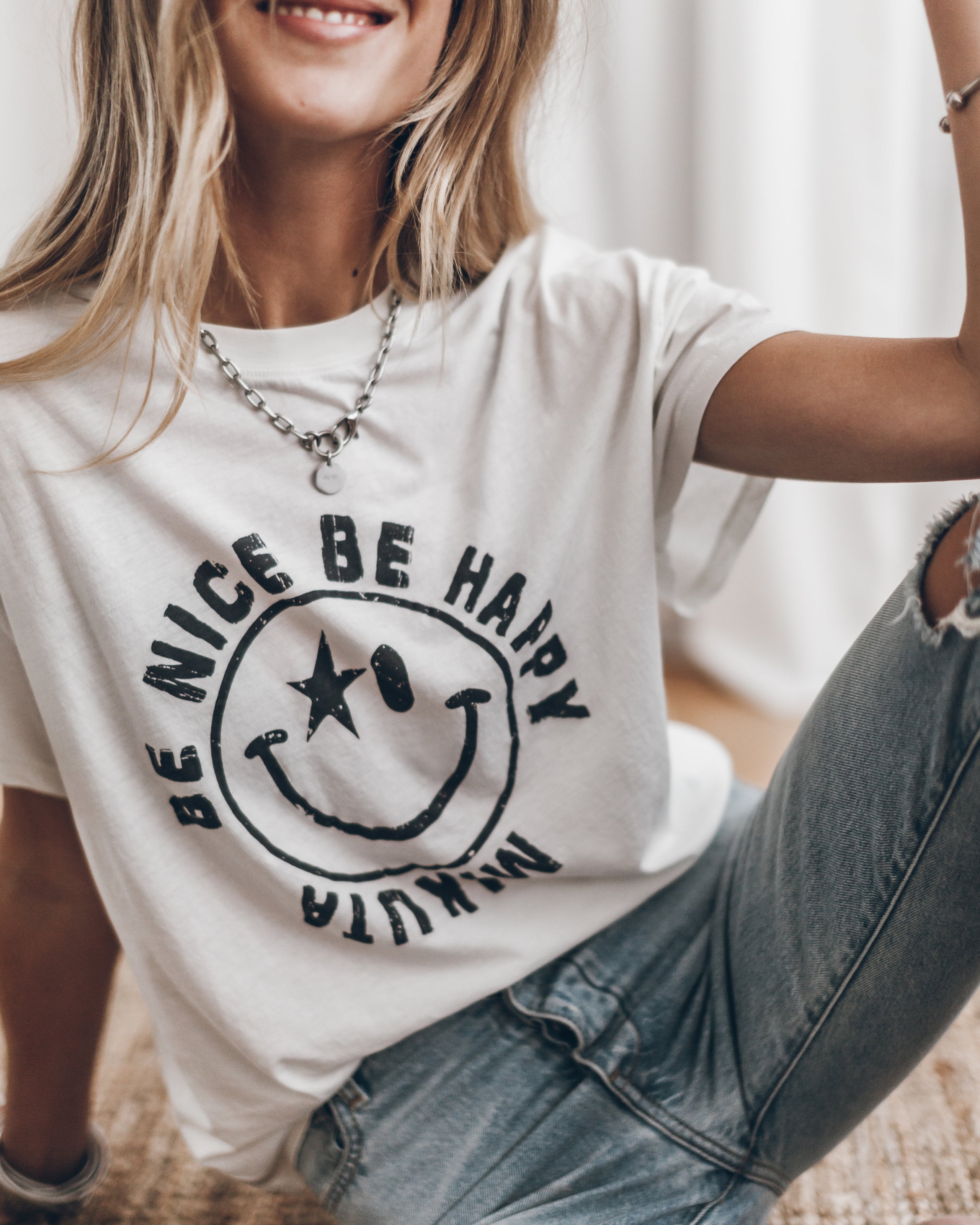 White cotton T-shirt with black smiley face and "Be Nice Be Happy" text, worn with blue jeans.