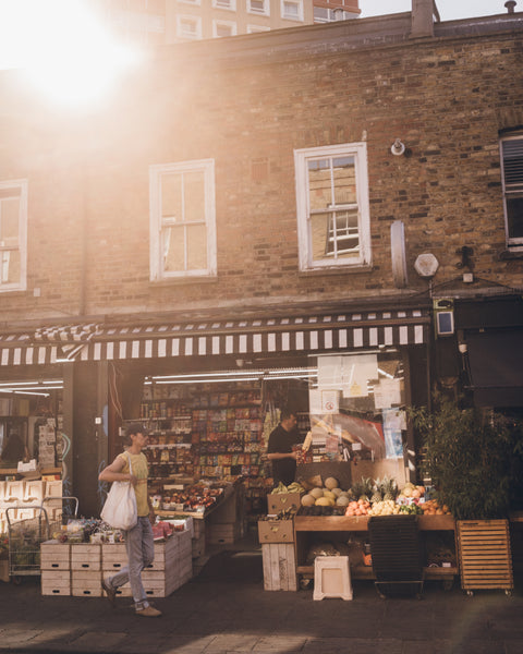 A warm, sunlit street scene outside a brick market shop with a striped awning, produce displays on the sidewalk, and a person carrying a white tote bag walking past.