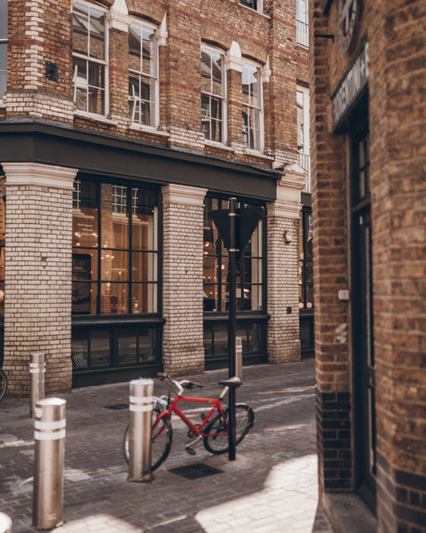 A red bicycle is parked against a black bollard lock on a cobblestone street in front of a brick and stone building with large black-framed windows.