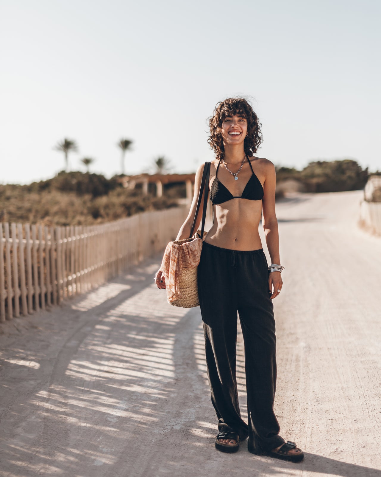 Woman in black bikini top and pants standing on a sandy path with a beach background