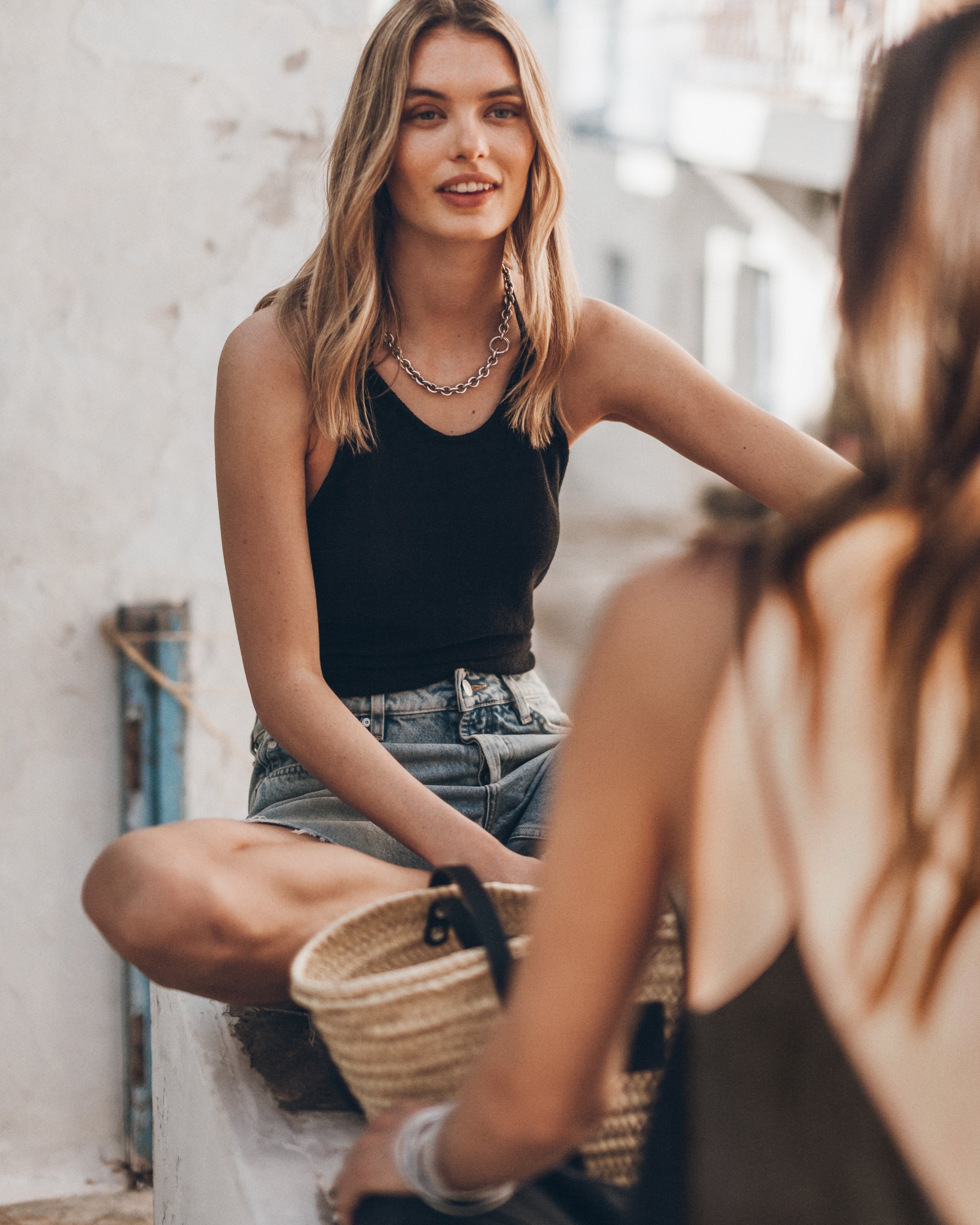 Woman sitting with a woven basket, wearing a black tank top and denim shorts.