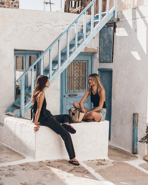 Two women sitting on a white ledge in a sunlit outdoor setting, with a straw bag between them.