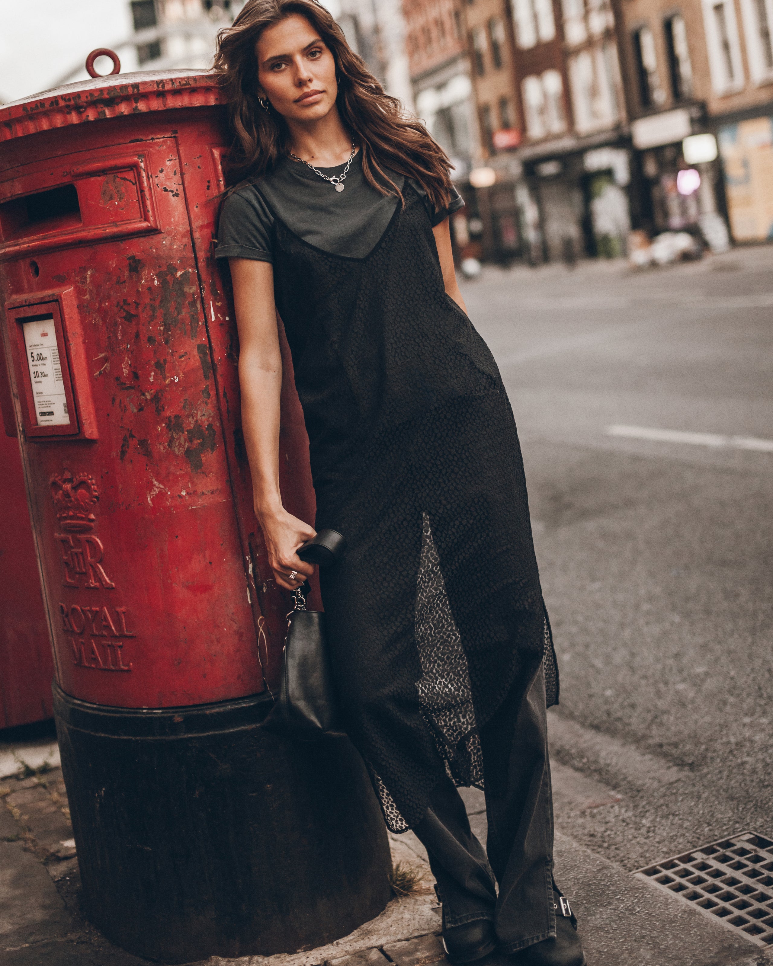 A woman stands on a city street wearing a black short-sleeve top and a long black lace dress.