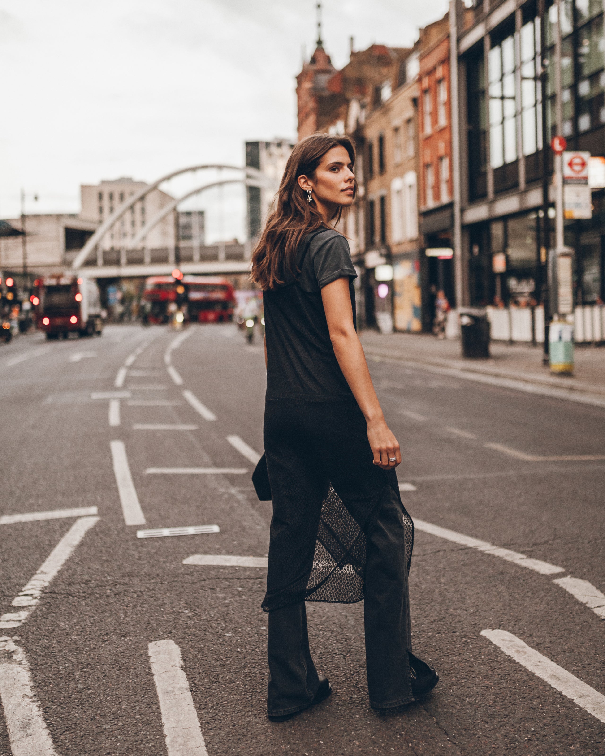 A woman stands on a city street wearing a dark gray short-sleeve t-shirt and black wide-leg trousers, with a black mesh bag hanging from her arm.