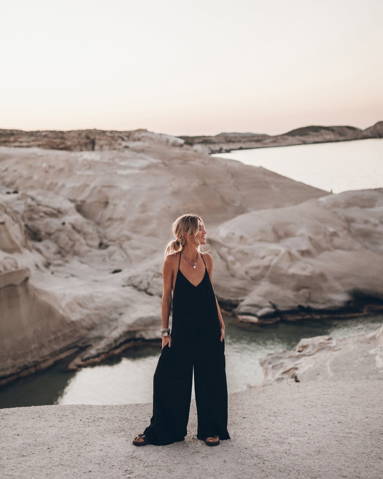 Woman in a black jumpsuit standing in front of a natural landscape with water and cliffs.