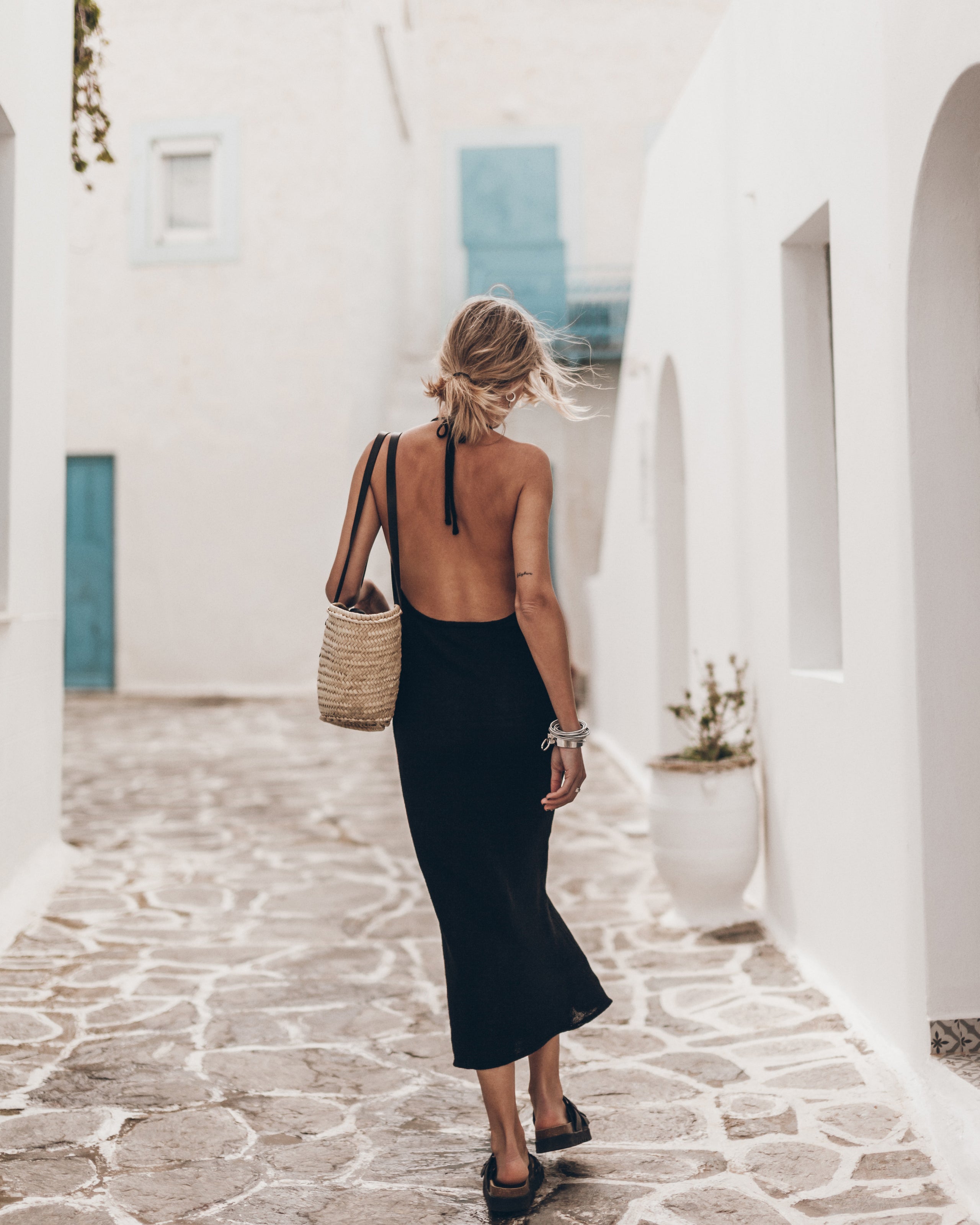 Woman in a black dress and a straw bag walking through a white-walled alleyway with blue doors.