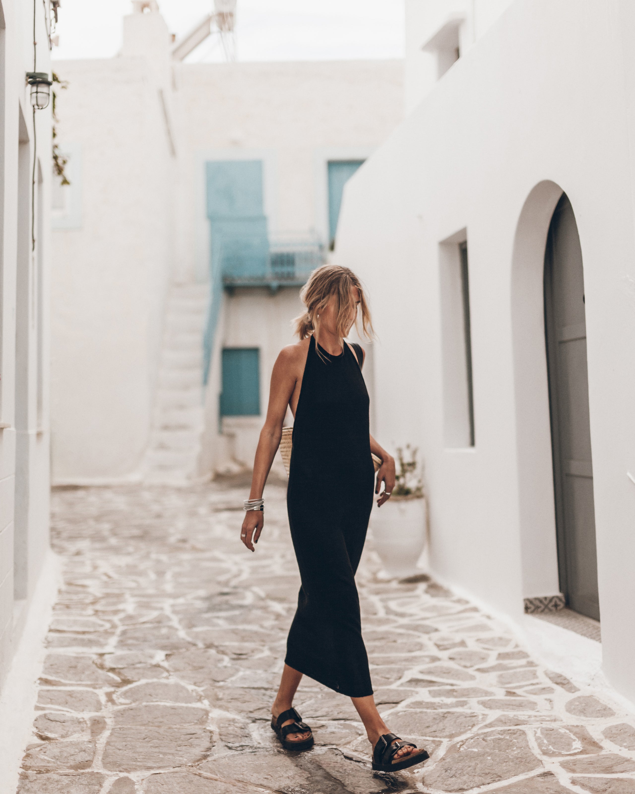 Woman in a black dress walking through a white-walled alleyway with stone pavement.