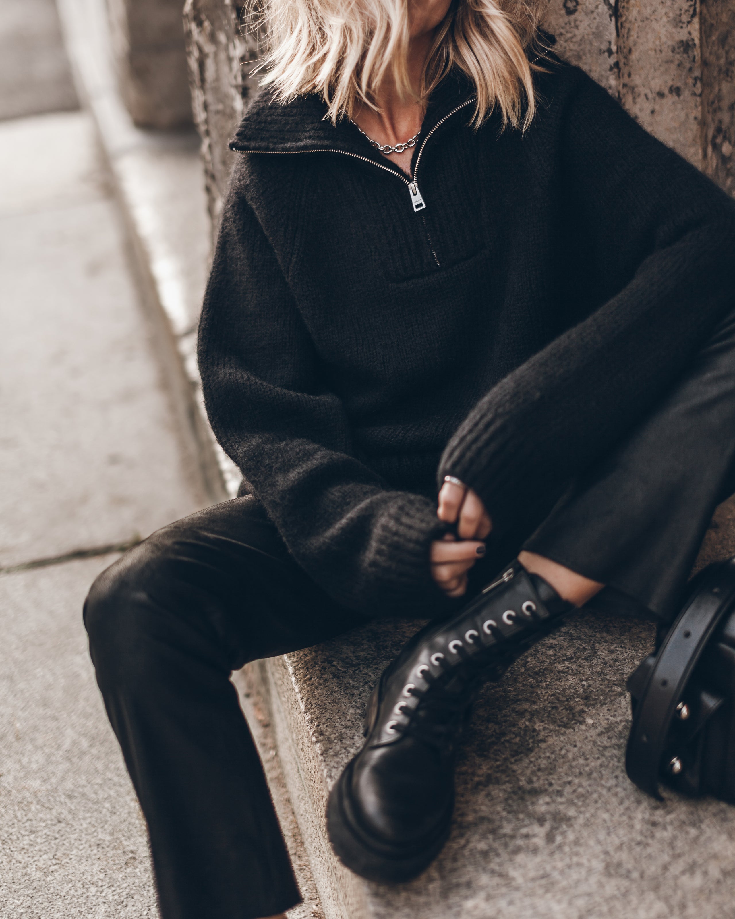 Person wearing a black outfit with a half-zip sweater and black boots sitting on a concrete surface.