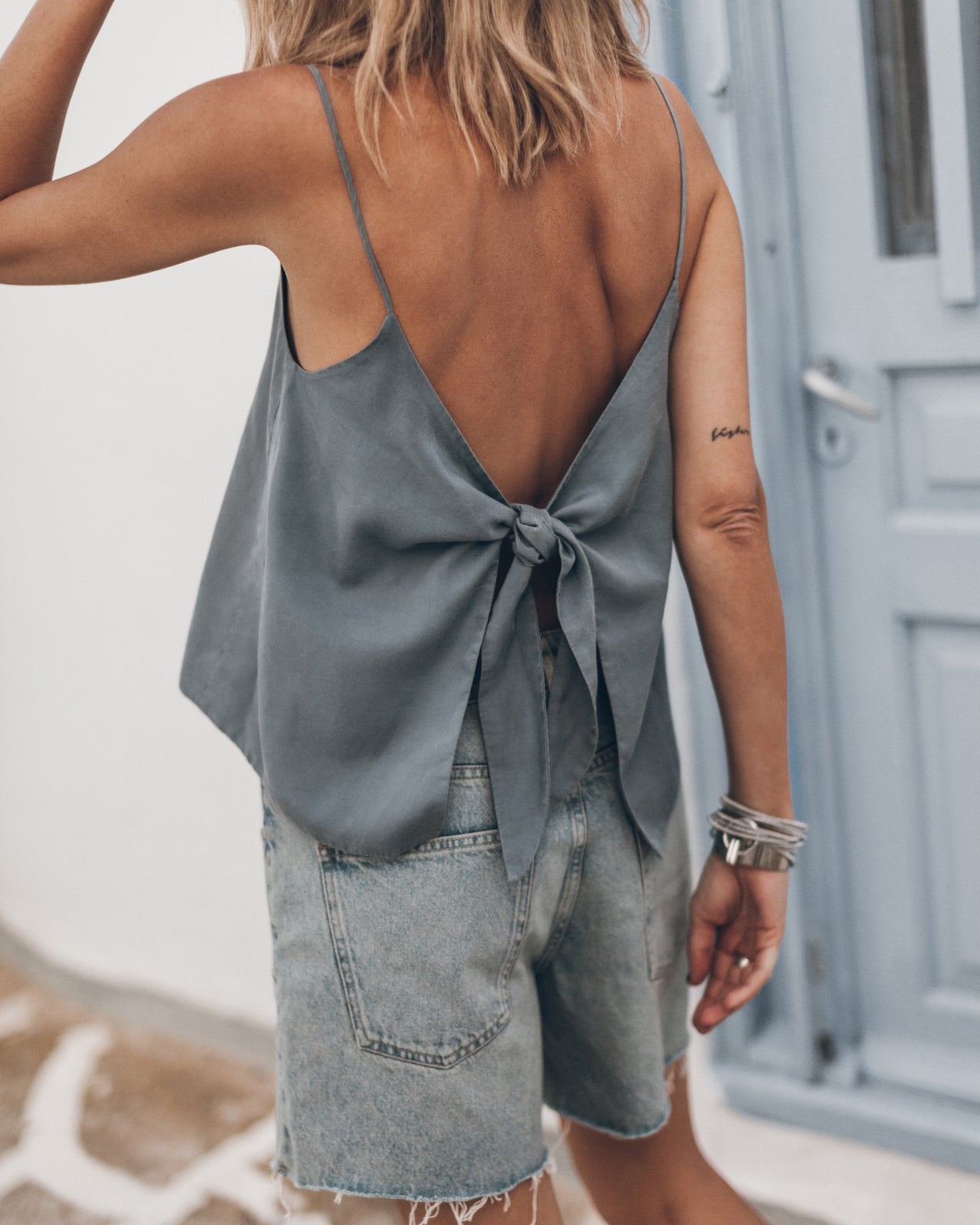Woman wearing a light blue sleeveless top with open back and denim shorts, standing in front of a door.