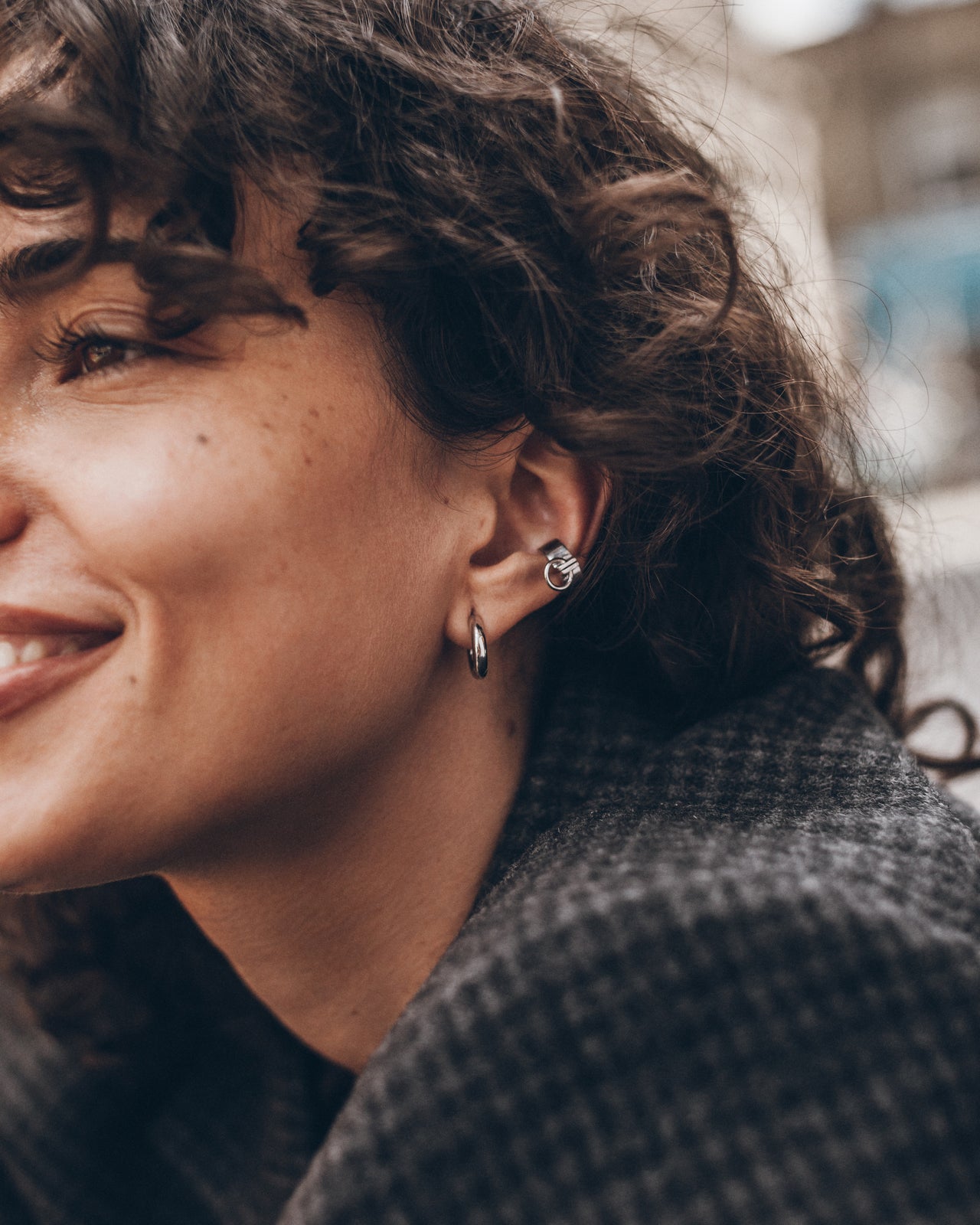 A close-up lifestyle shot of a woman with curly brown hair wearing a small silver hoop earring and a matching silver stud earring.