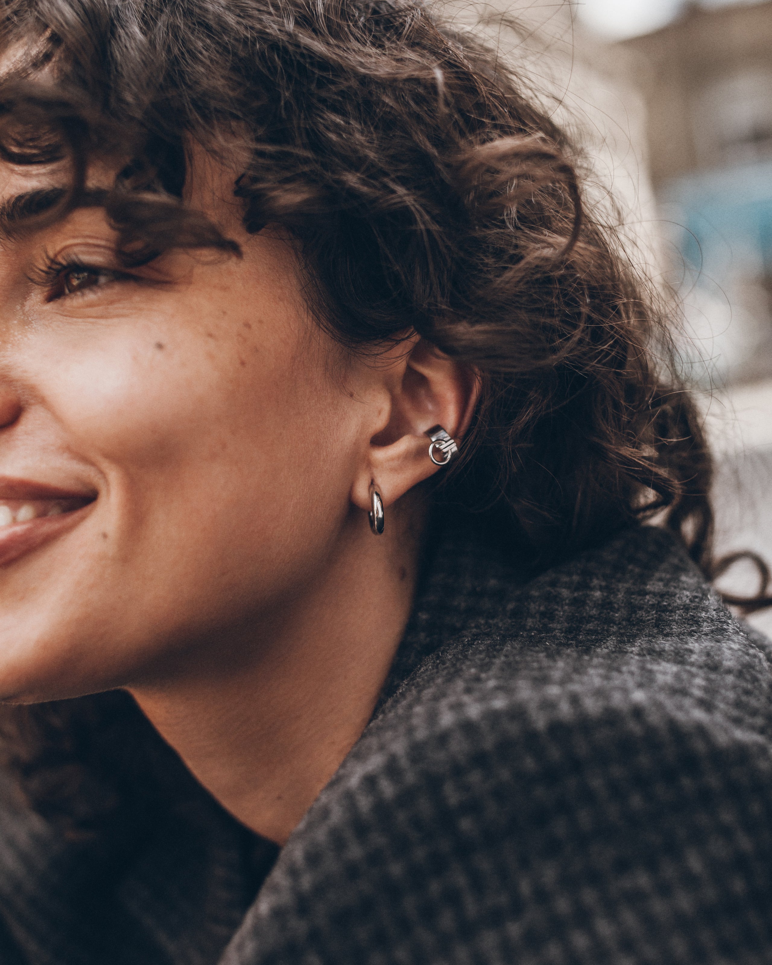 A close-up lifestyle shot of a woman with curly brown hair wearing a small silver hoop earring and a matching silver stud earring.