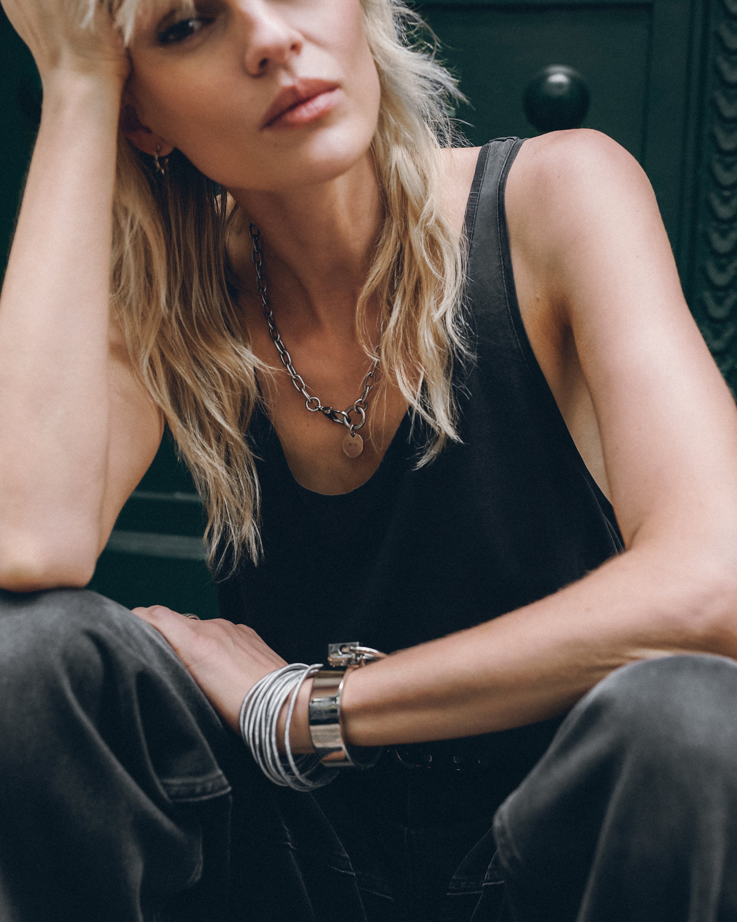 Woman wearing a black sleeveless top and dark pants with jewelry, sitting against a dark background.
