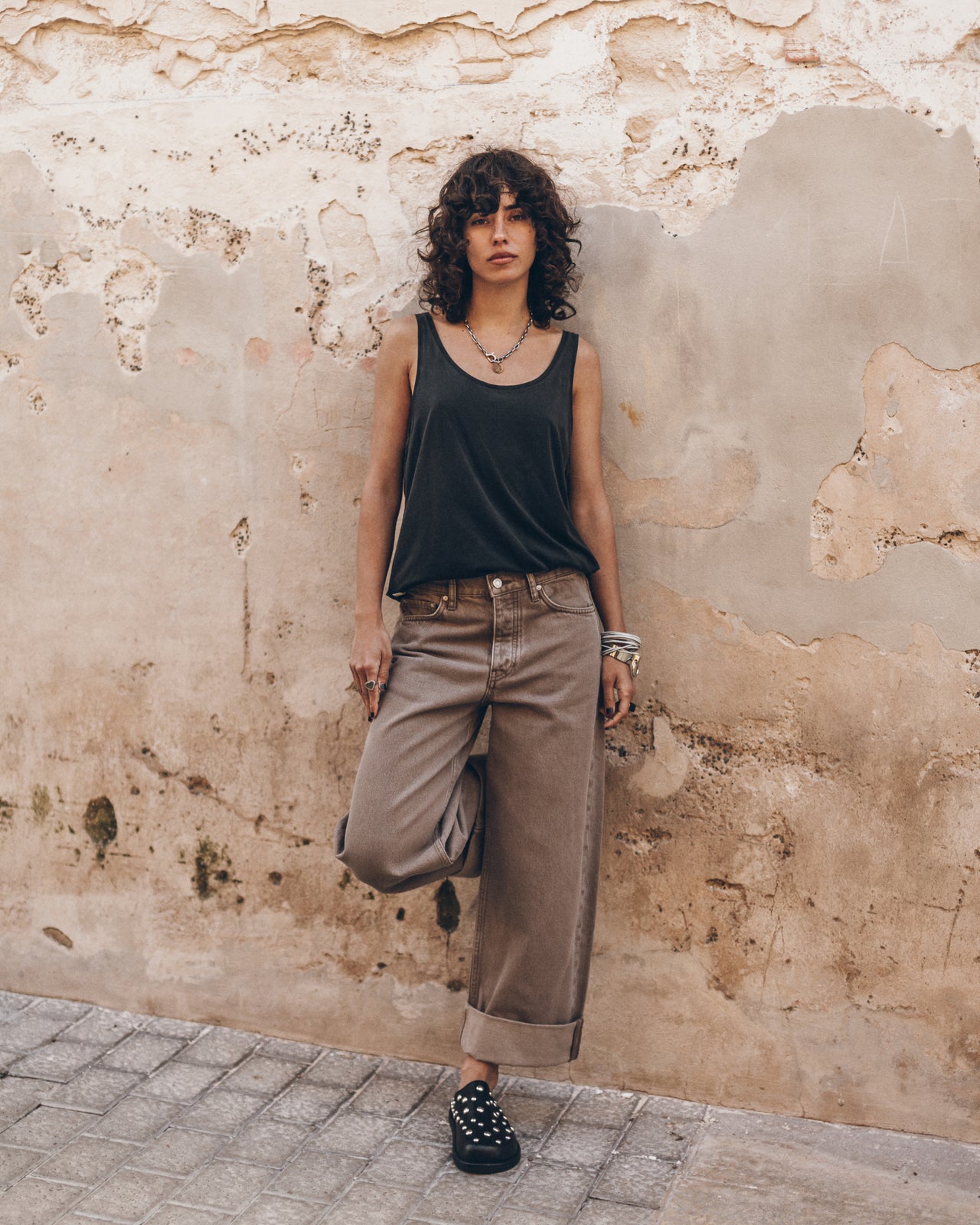 Woman wearing a black tank top and chestnut wash jeans standing against a textured wall.