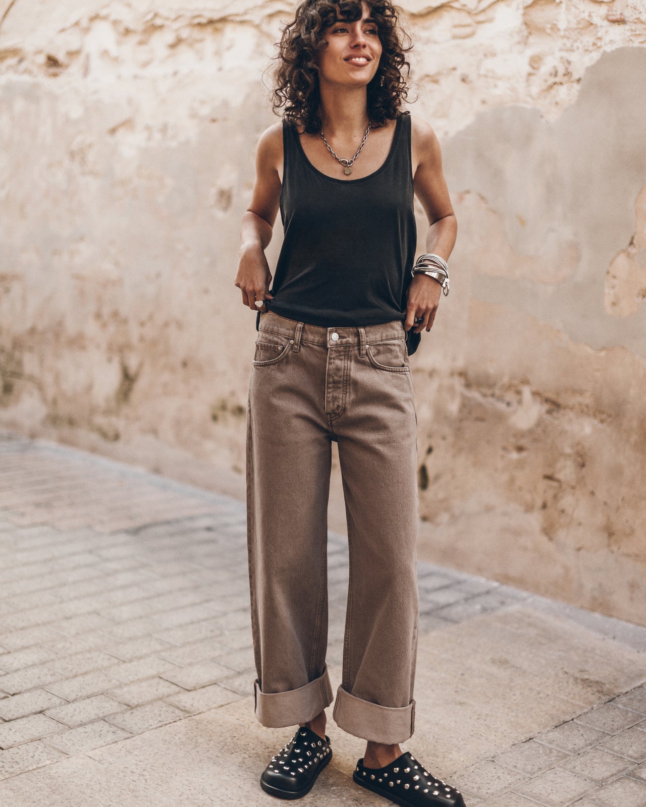 Woman wearing a black tank top and chestnut wash jeans standing against a textured wall.
