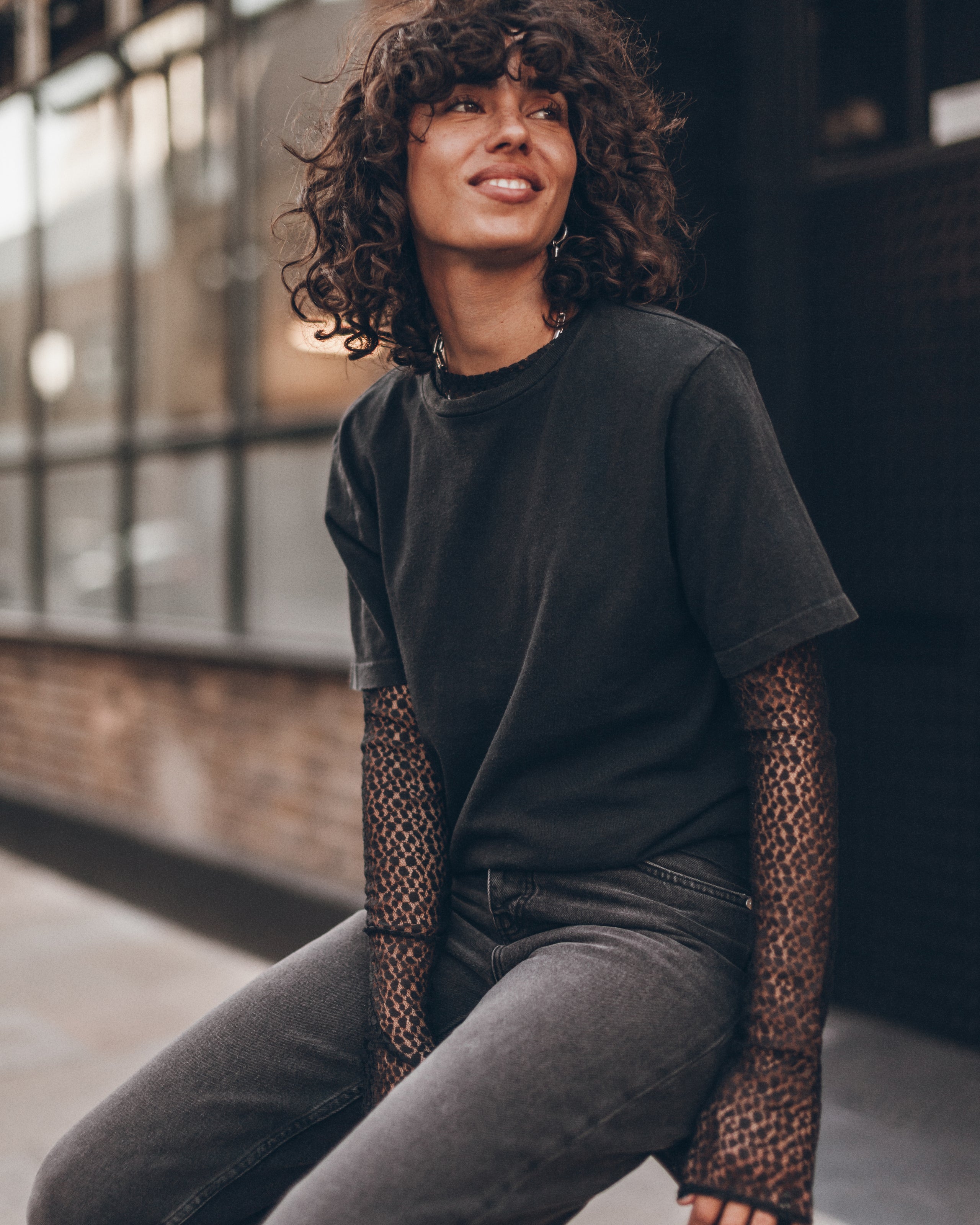 A woman with curly dark hair wearing a black crewneck t-shirt with sheer black lace long sleeves, paired with gray jeans, sitting in an urban setting.