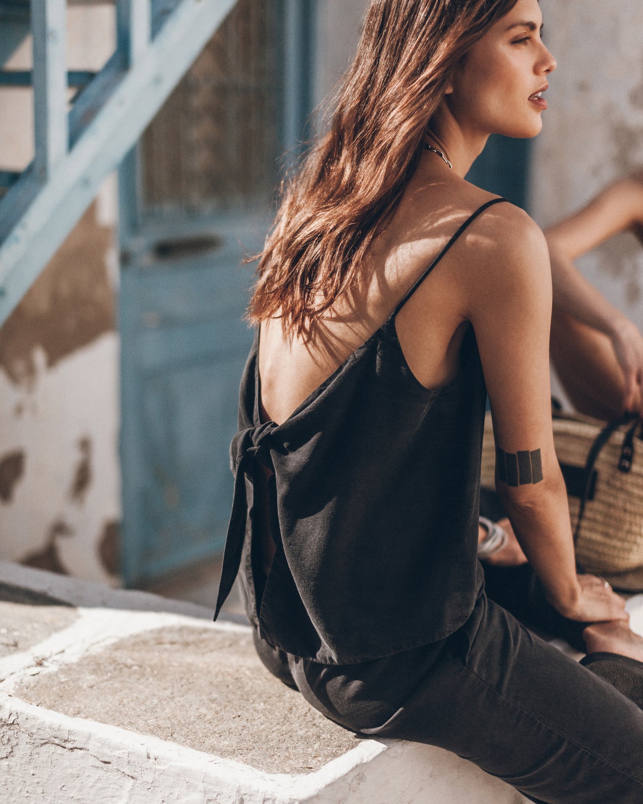 Woman sitting outdoors wearing a black sleeveless top and pants, with a woven bag in the background.