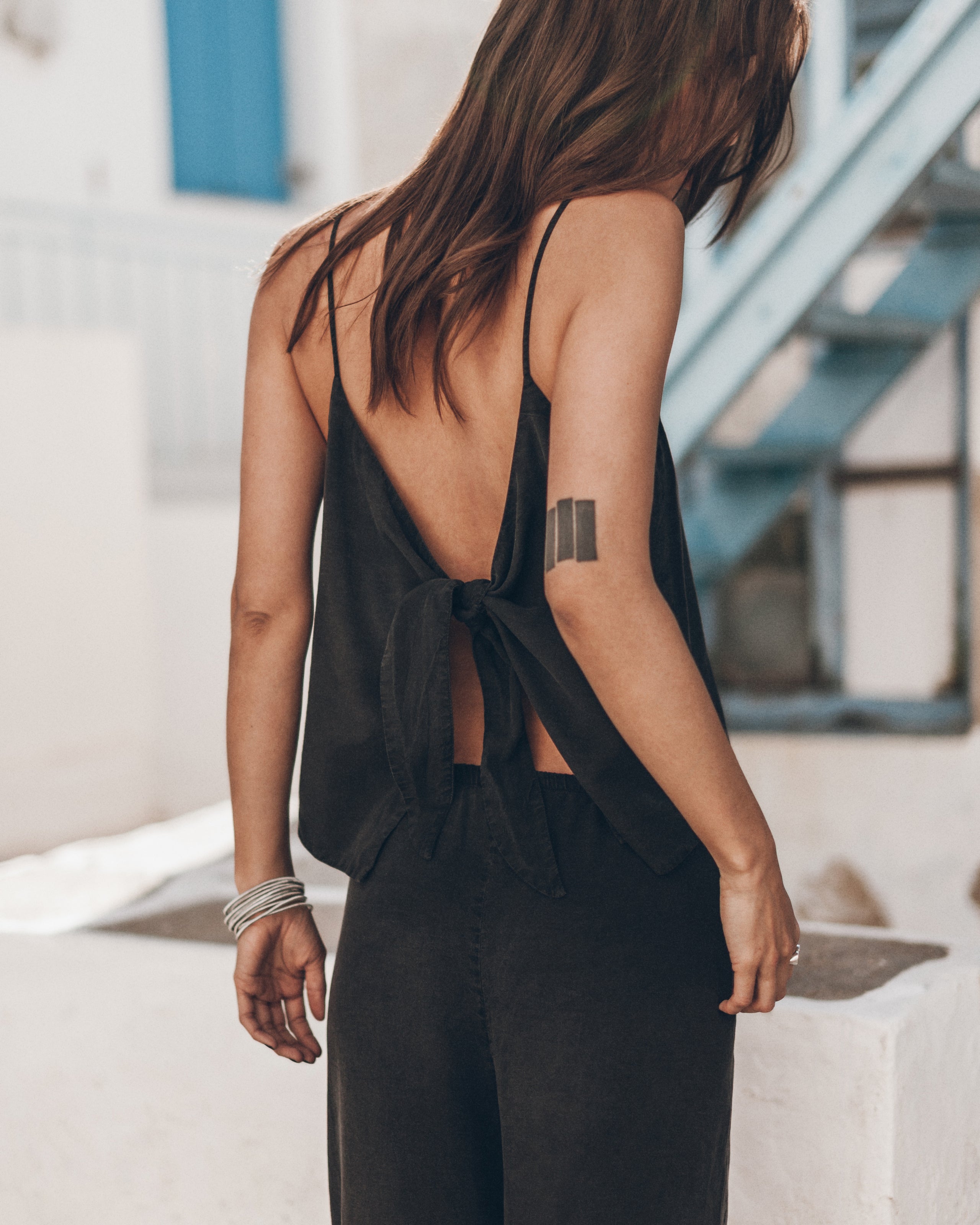 Woman wearing a black open-back top and black pants with a tattoo on their arm, standing against a white wall.