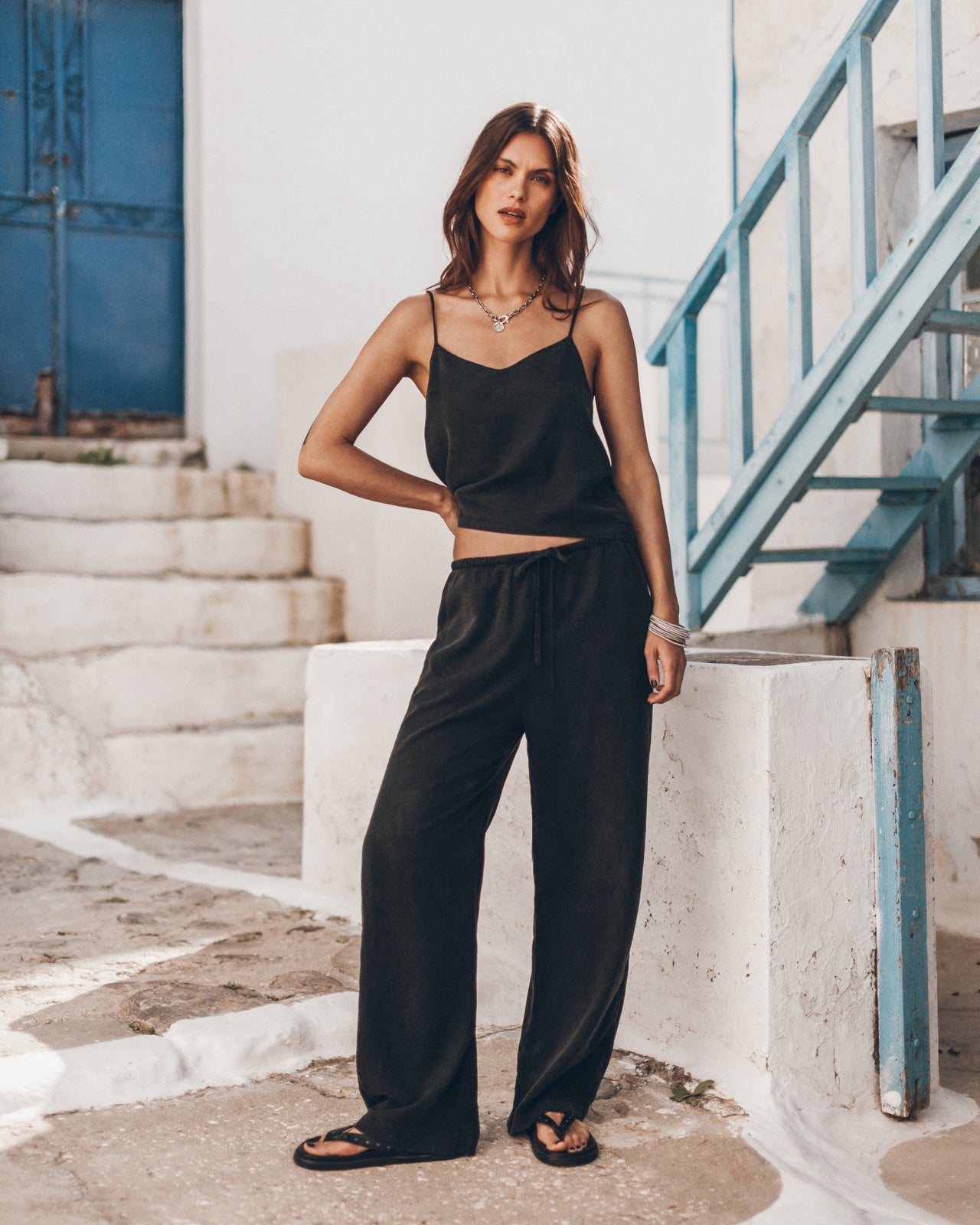 Woman in a black outfit wearing a black top and comfortable black pants, standing on a stone path with a blue metal staircase in the background.