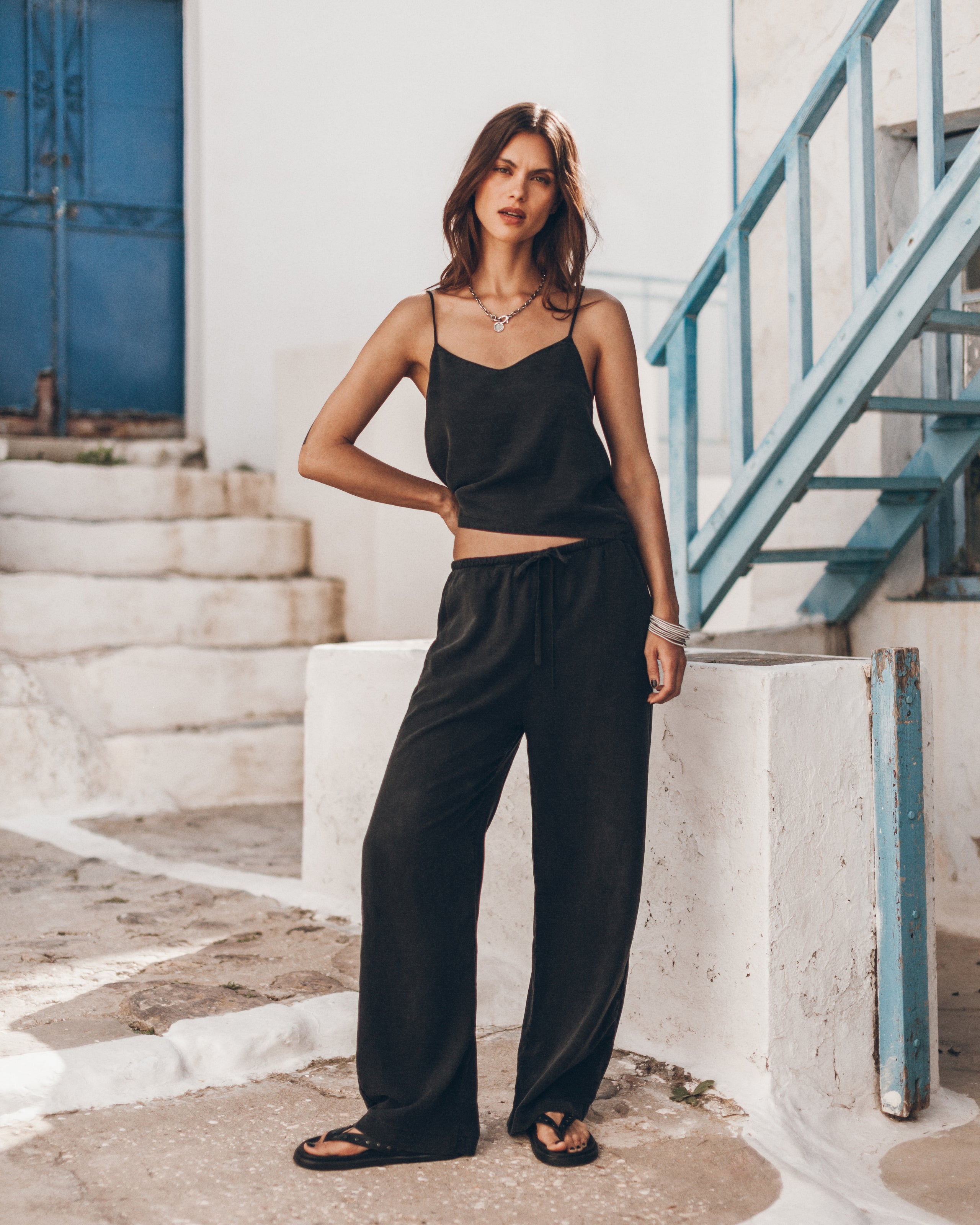 Woman in a black outfit wearing a black top and comfortable black pants, standing on a stone path with a blue metal staircase in the background.