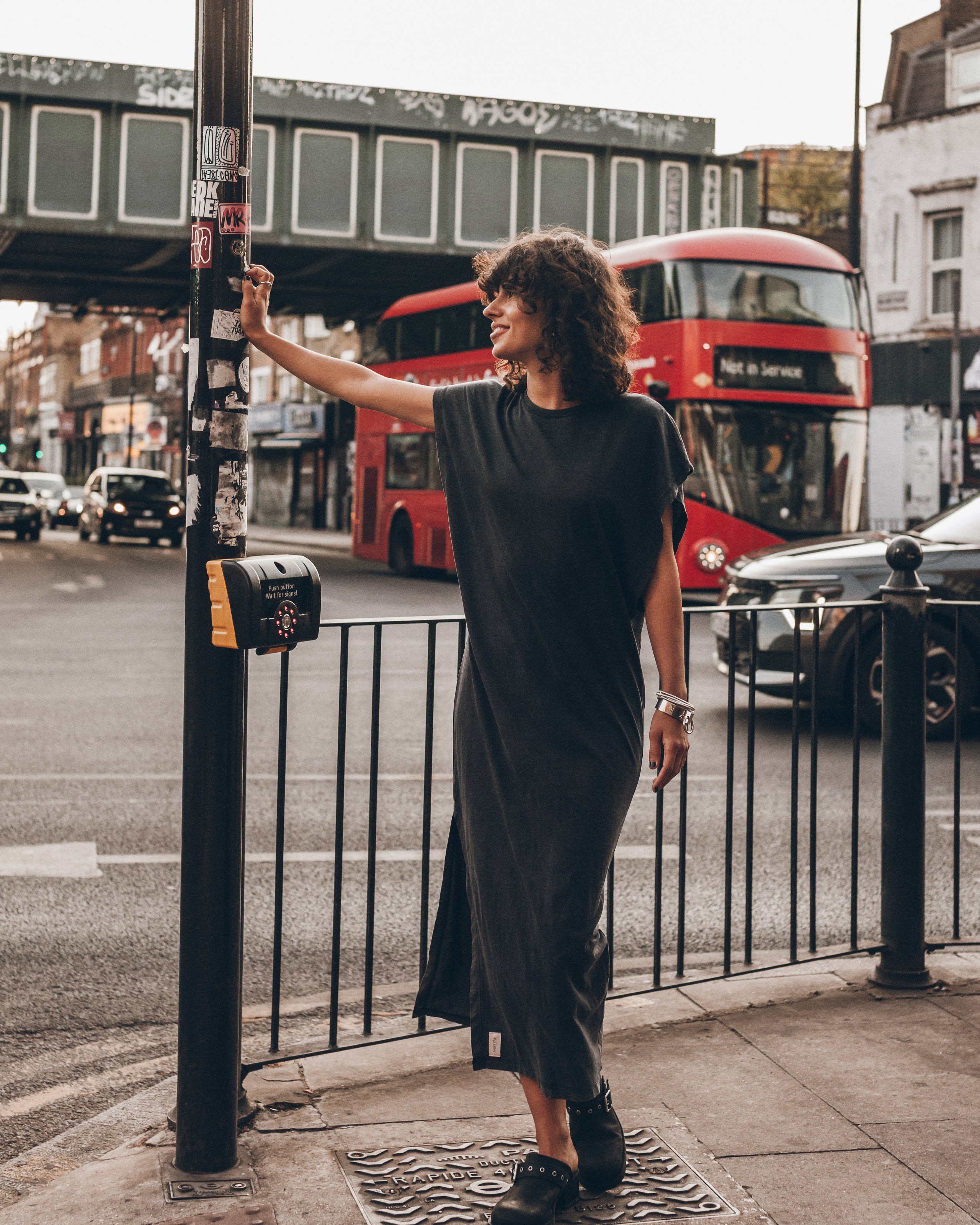 A woman wearing a black midi-length sleeveless dress and black loafers stands on a city sidewalk next to a pole, with a red double-decker bus and urban street scene in the background.