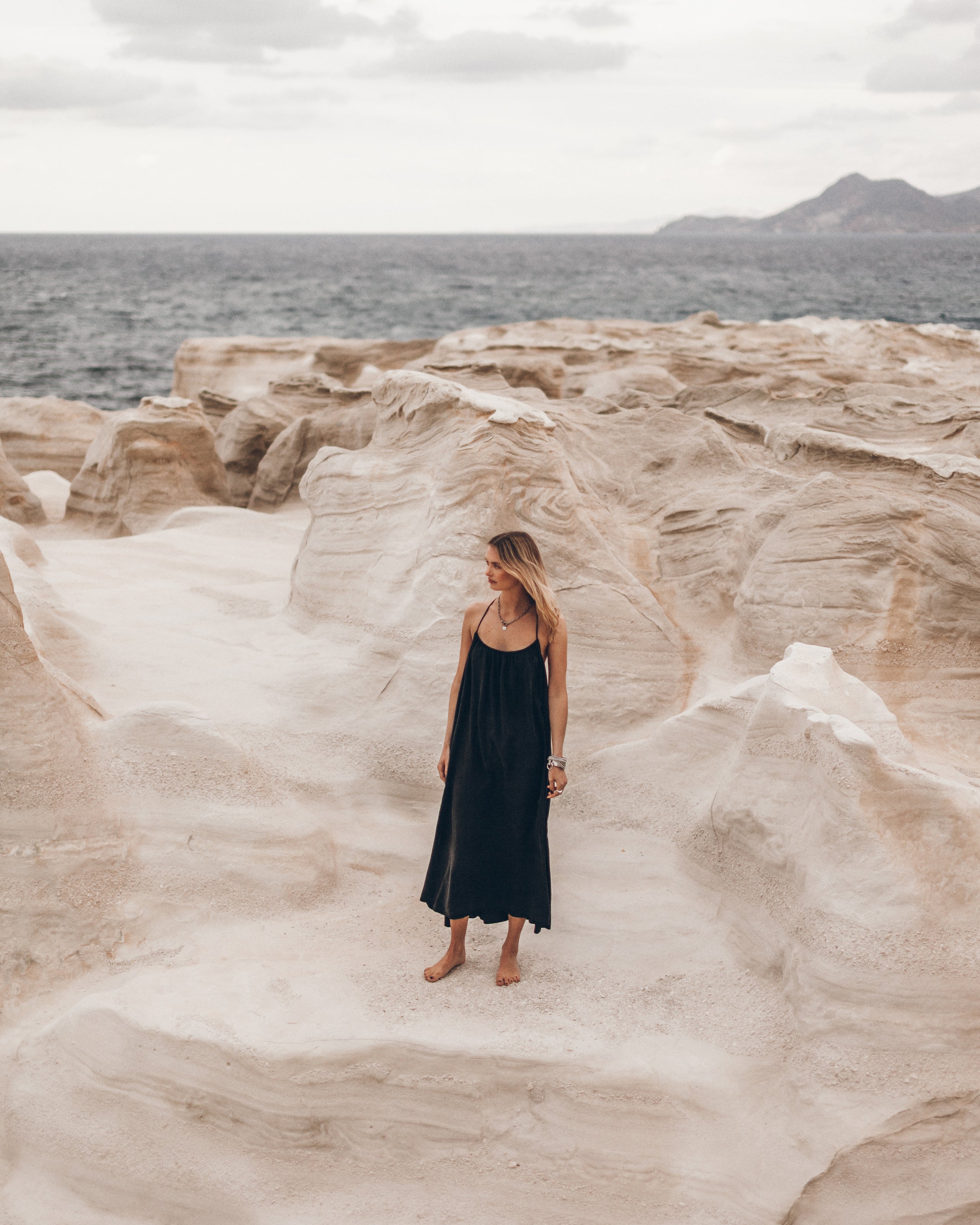 Woman in a long black dress standing on a rocky coastal landscape with ocean and mountains in the background.