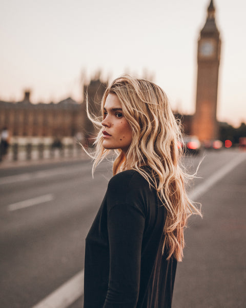 A woman stands on a street in London, wearing a black long-sleeve dress. The background shows a softly blurred Big Ben and Houses of Parliament at sunset.