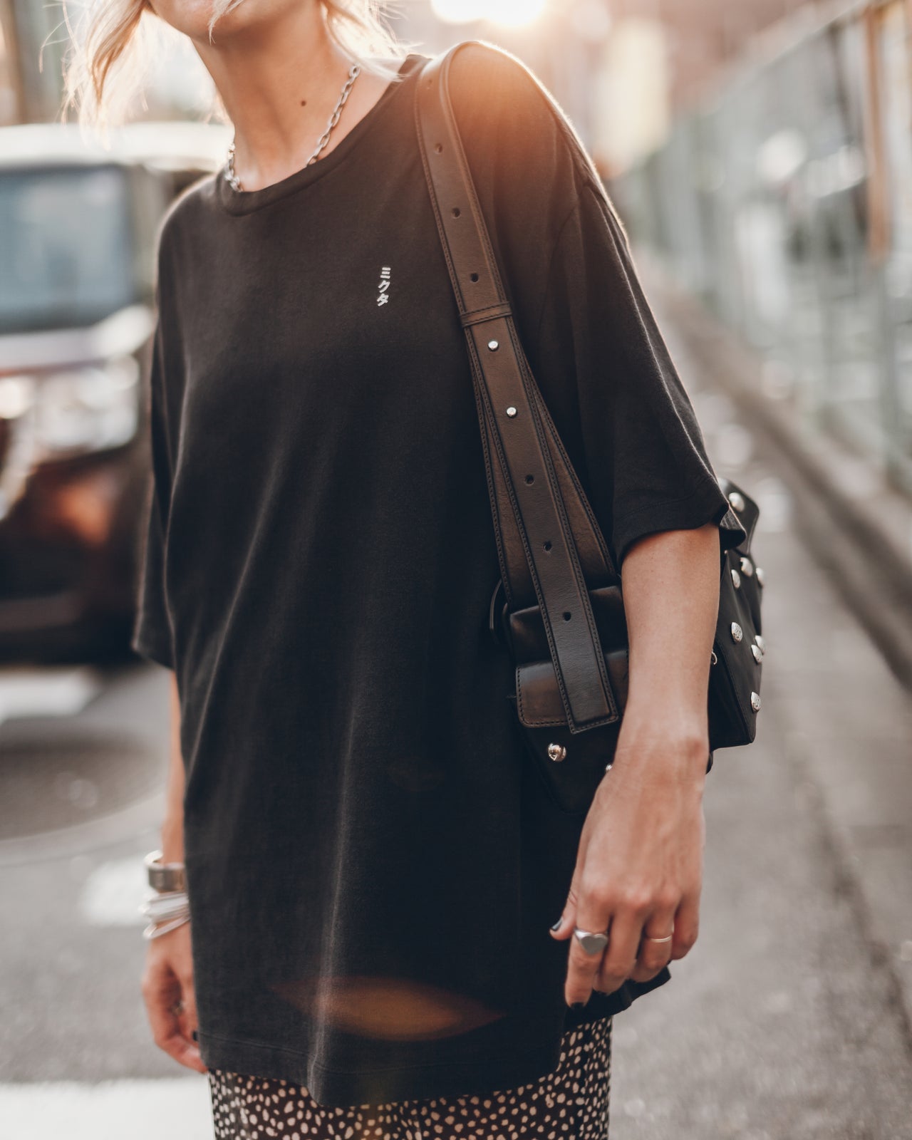 Person wearing a black oversized shirt with a brand logo, carrying a brown leather bag on a street.