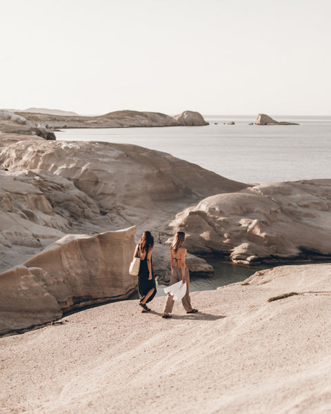 Two people walking on a rocky beach with a body of water and distant landforms.