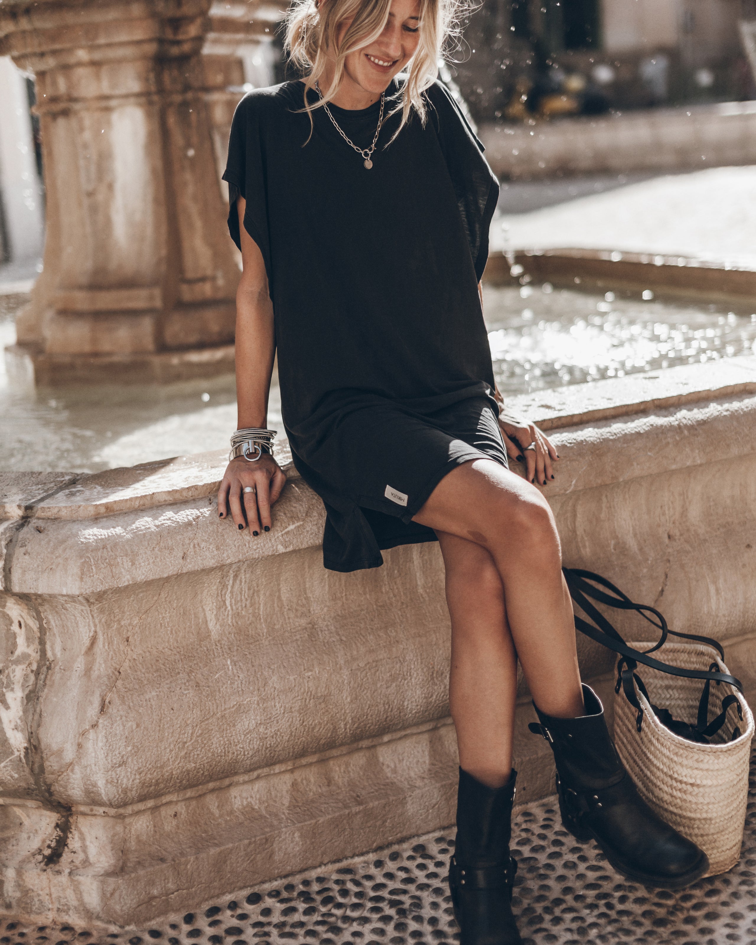 A woman wearing a black oversized short-sleeve t-shirt dress and black shorts sits on a stone fountain ledge in an outdoor setting. She is wearing black ankle boots and has a woven straw handbag next to her.