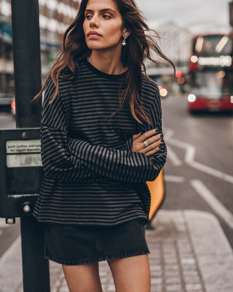 A woman stands on a city street wearing a black and gray striped longsleeve top and a black denim skirt.