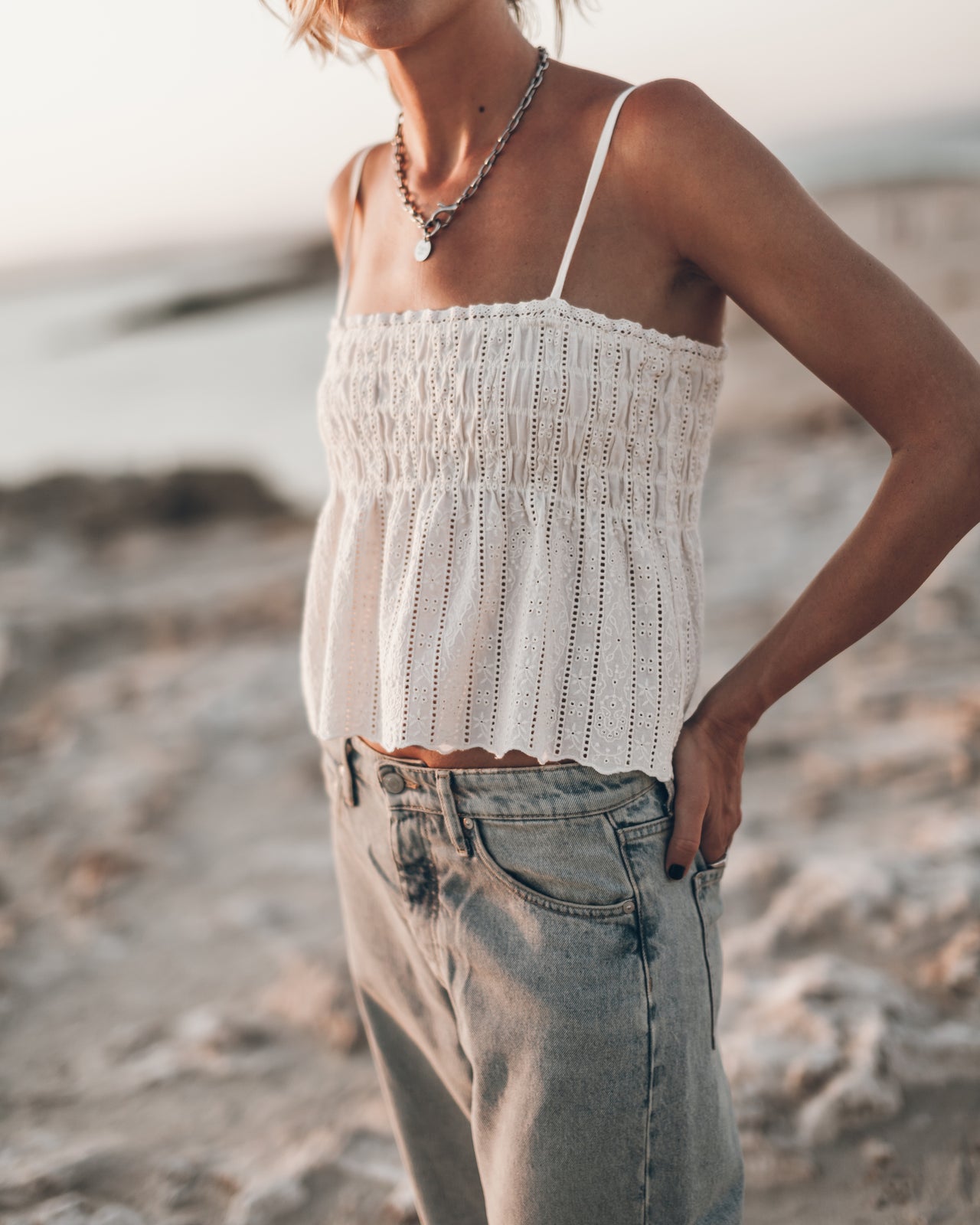 Person wearing a white crochet top and light blue jeans on a beach.