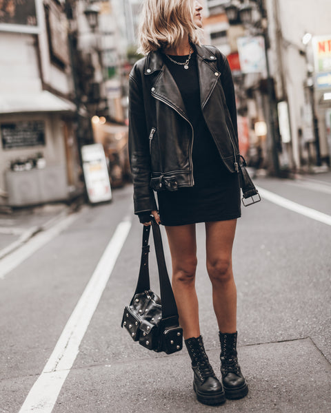 Person wearing a black leather jacket, black dress, and black boots on a city street.