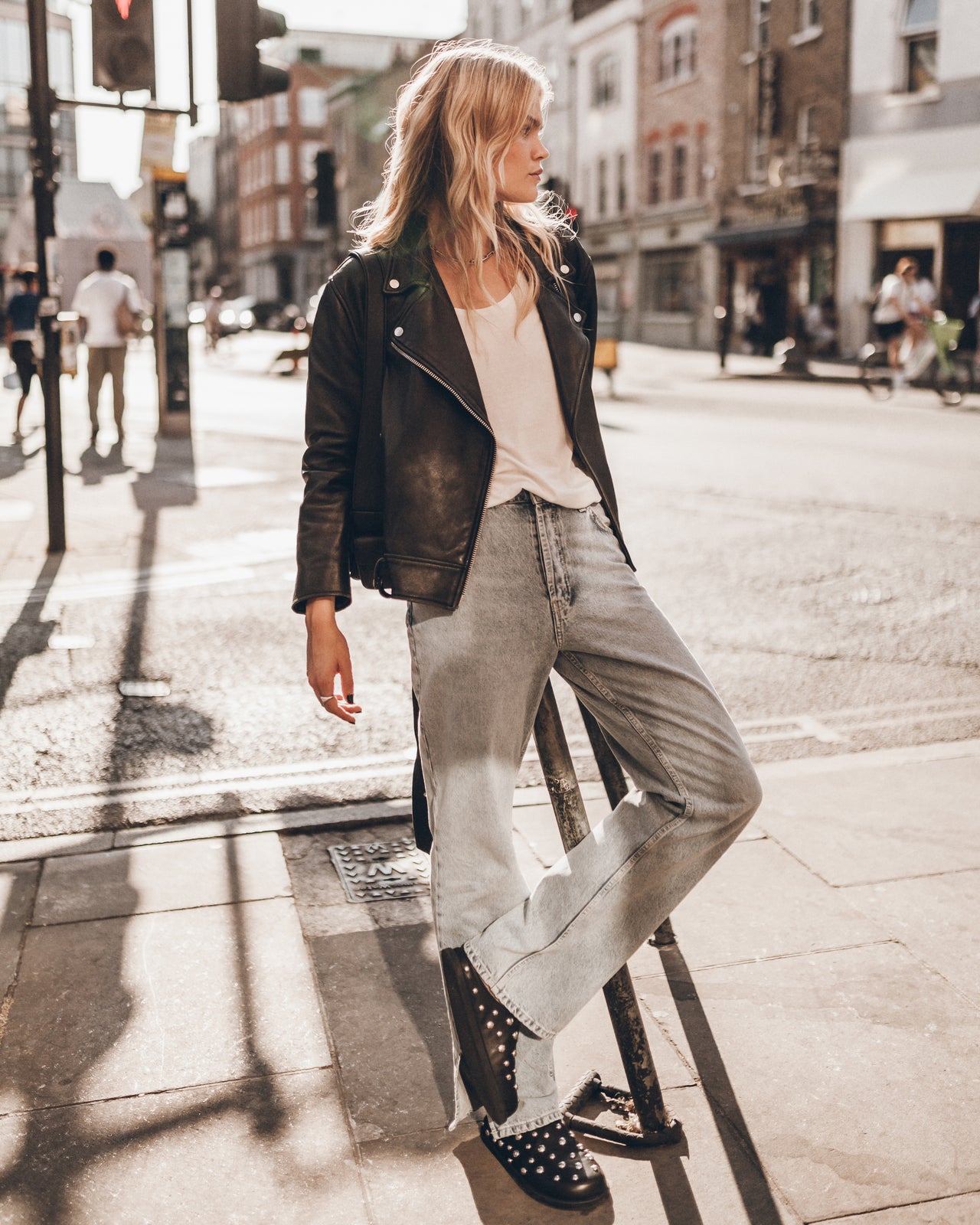 A woman stands on a city street corner wearing a black leather jacket, white tank top, light-wash wide-leg jeans, and black studded flat shoes.