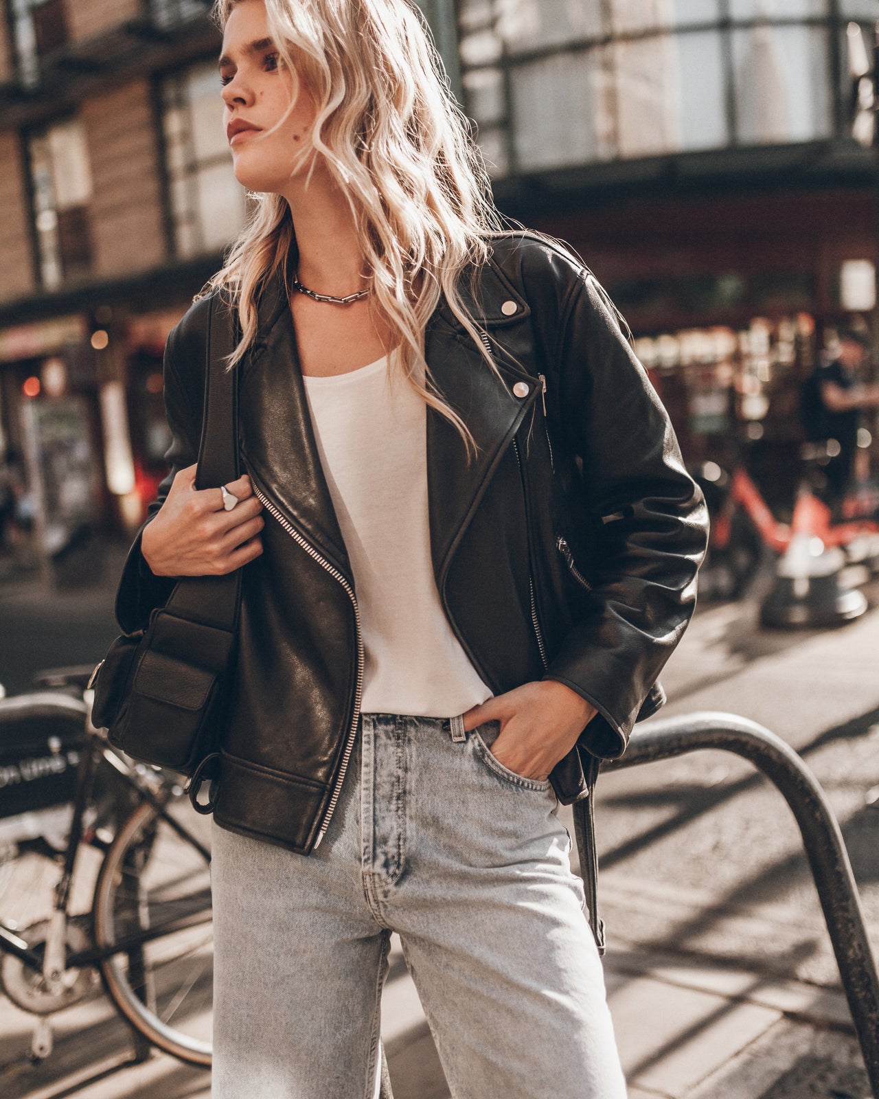 A woman wearing a black leather jacket, white tank top, and light wash jeans stands on a city street.