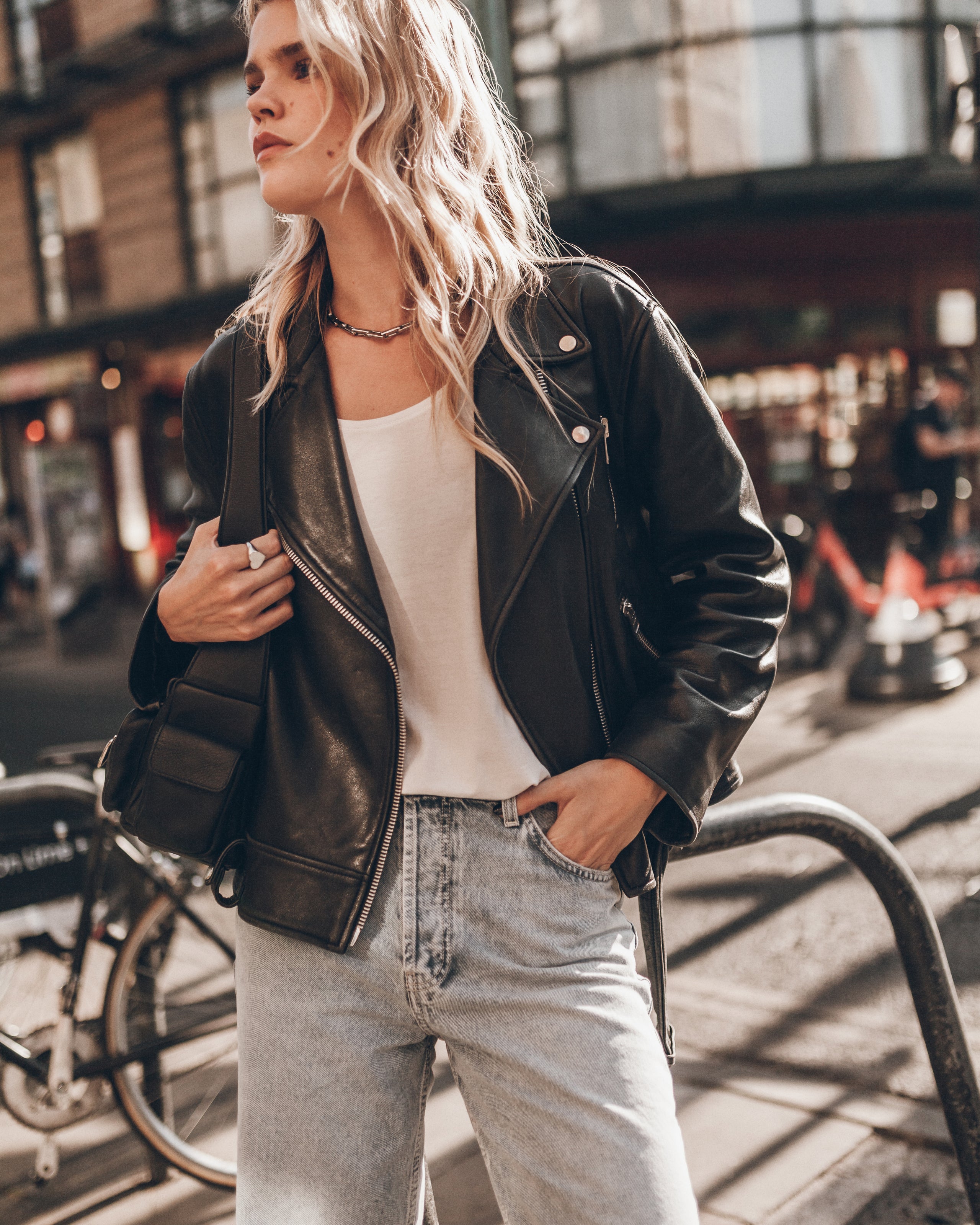 A woman wearing a black leather jacket, white tank top, and light wash jeans stands on a city street.