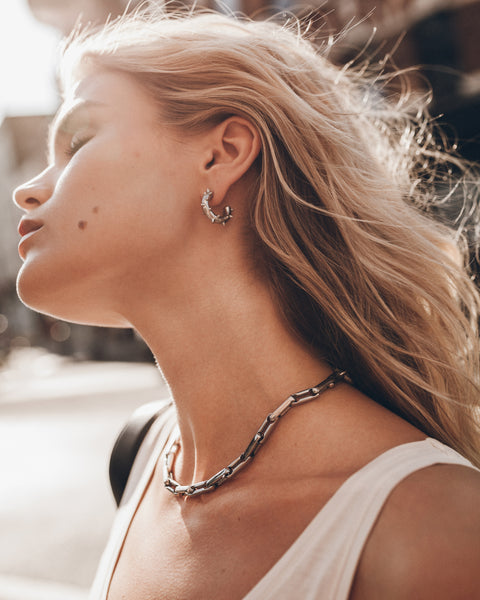 A close-up lifestyle shot of a woman wearing a chunky silver chain necklace and a matching silver crescent-shaped hoop earring.