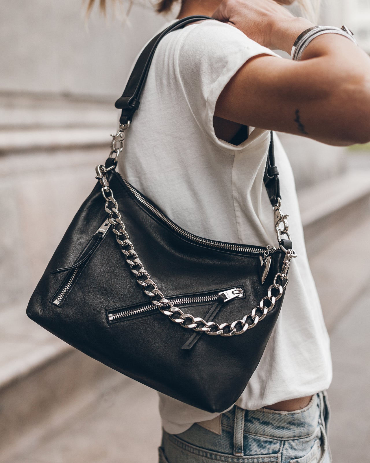 Woman holding a black handbag with a chain detail, wearing a white t-shirt and blue jeans.