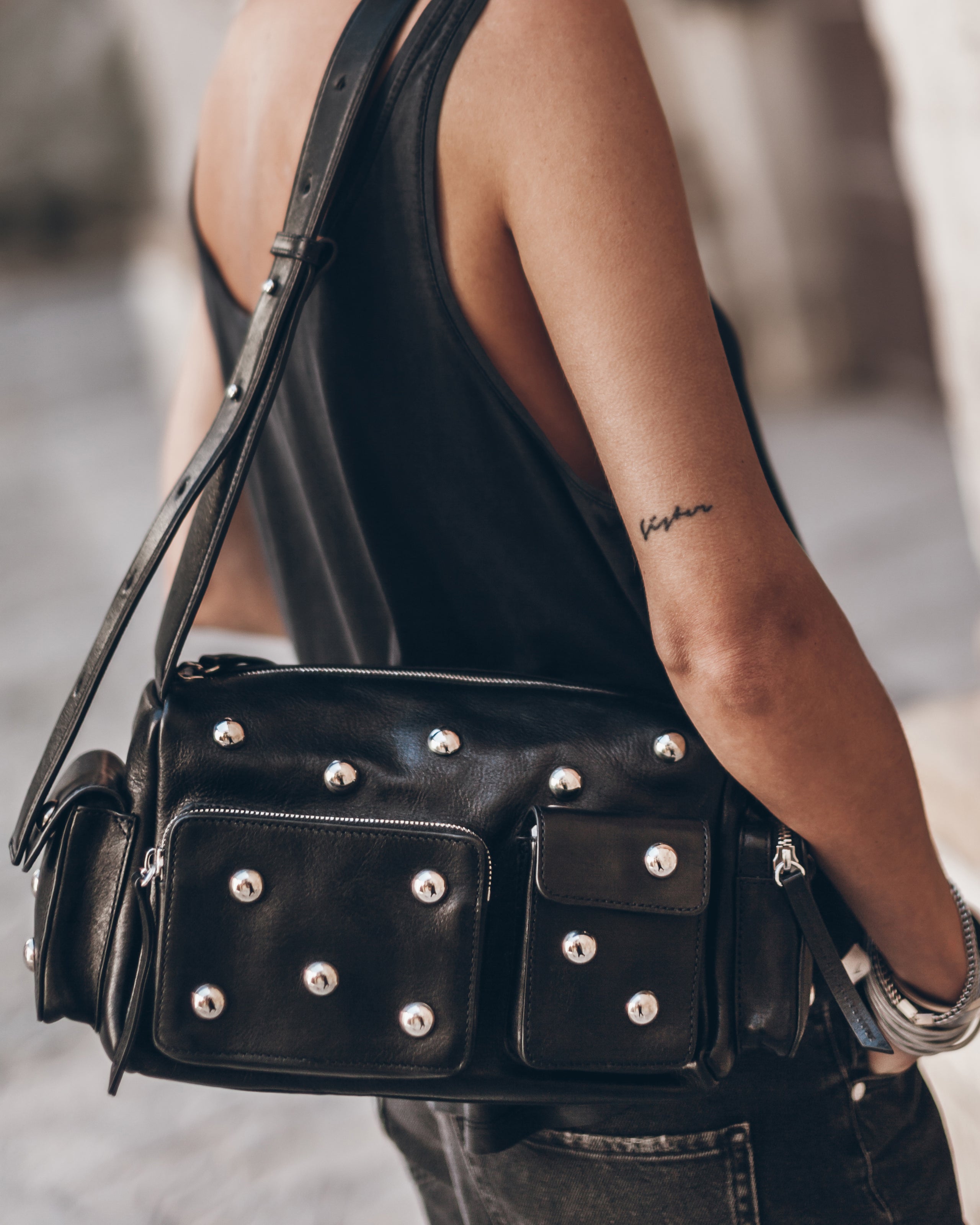 Woman in black tank top with studded black leather shoulder bag, silver bracelets, and arm tattoo.