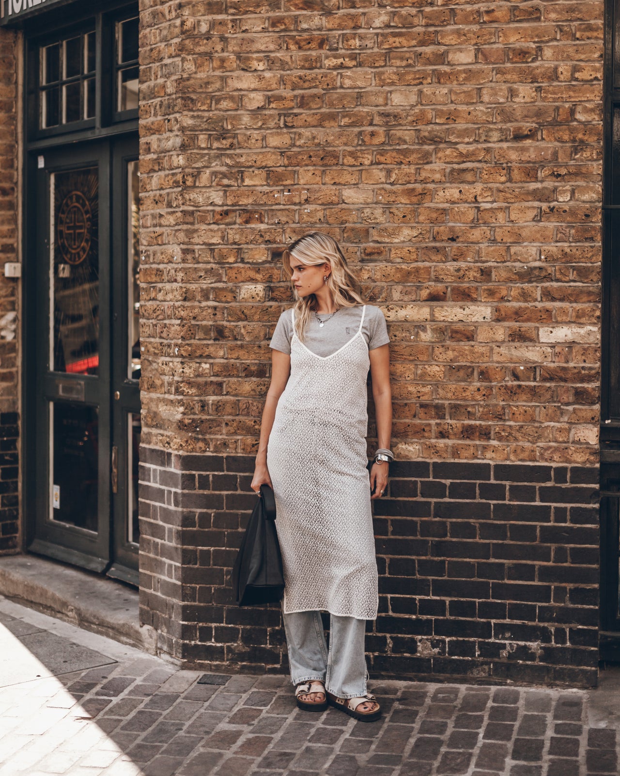 A woman stands on a brick sidewalk, wearing a light gray t-shirt under a white textured pinafore dress, light gray wide-leg pants, and brown sandals, while holding a black tote bag.