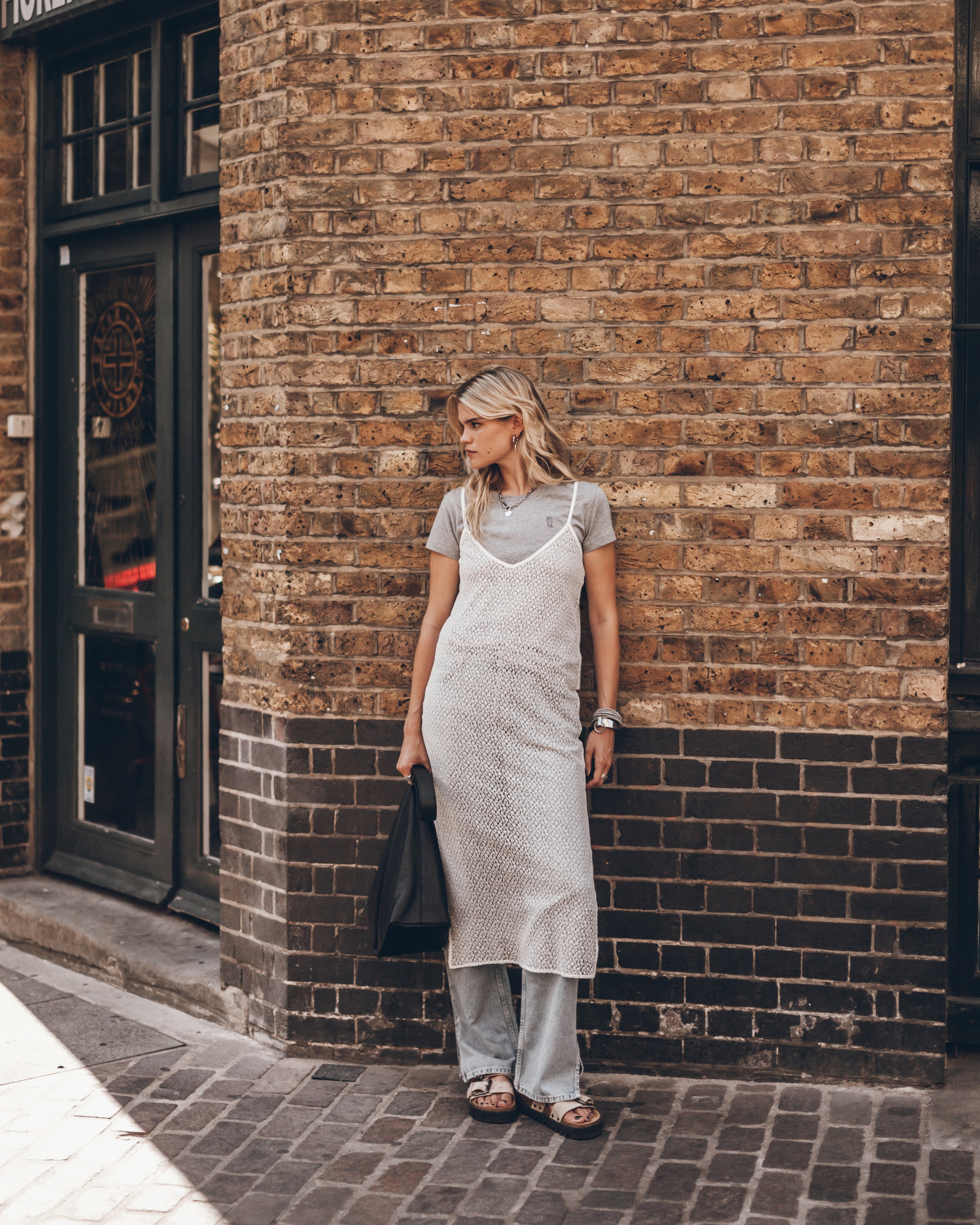 A woman stands on a brick sidewalk, wearing a light gray t-shirt under a white textured pinafore dress, light gray wide-leg pants, and brown sandals, while holding a black tote bag.
