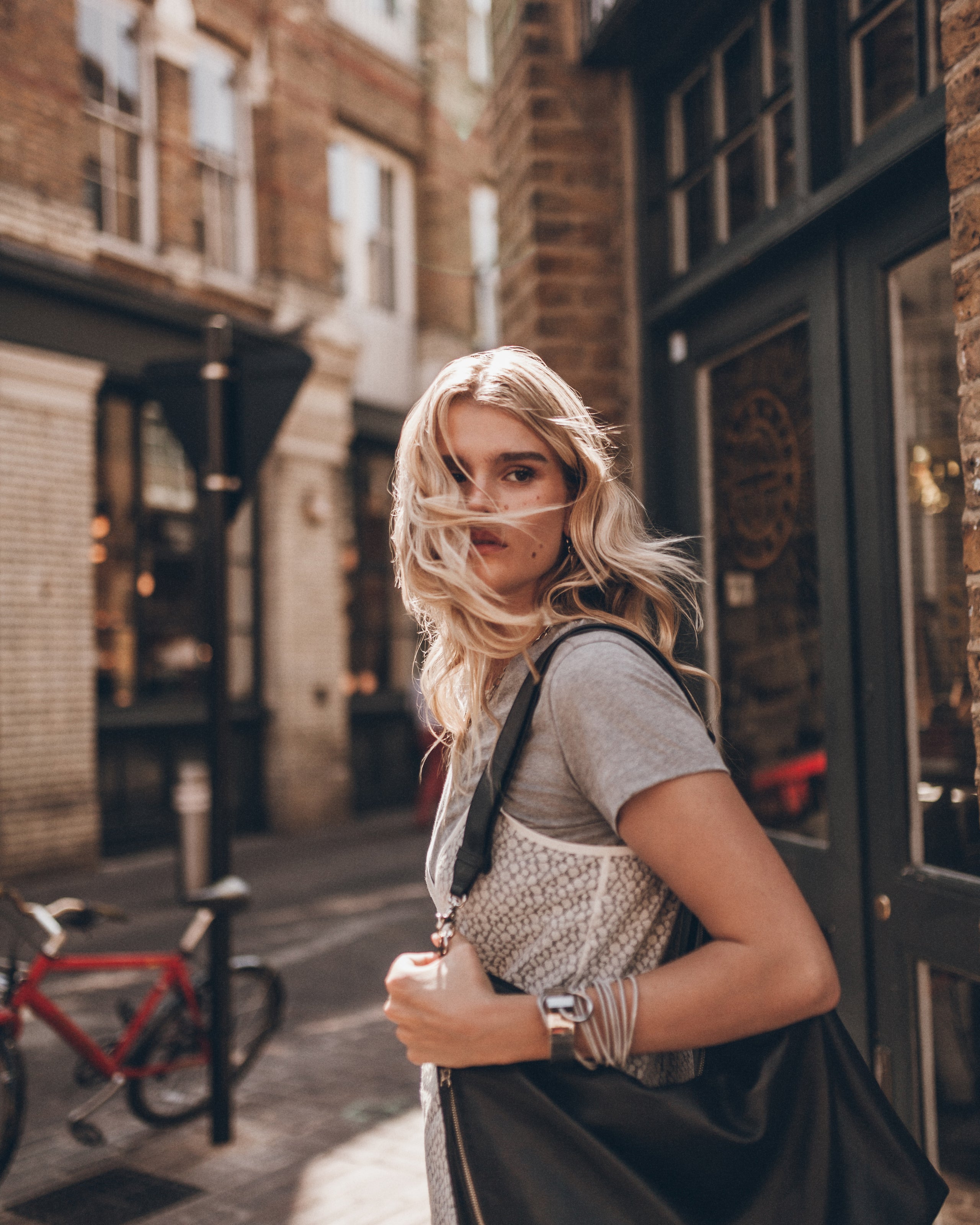 A blonde woman stands on a city street, wearing a gray t-shirt, a white knit dress, and carrying a large black shoulder bag.